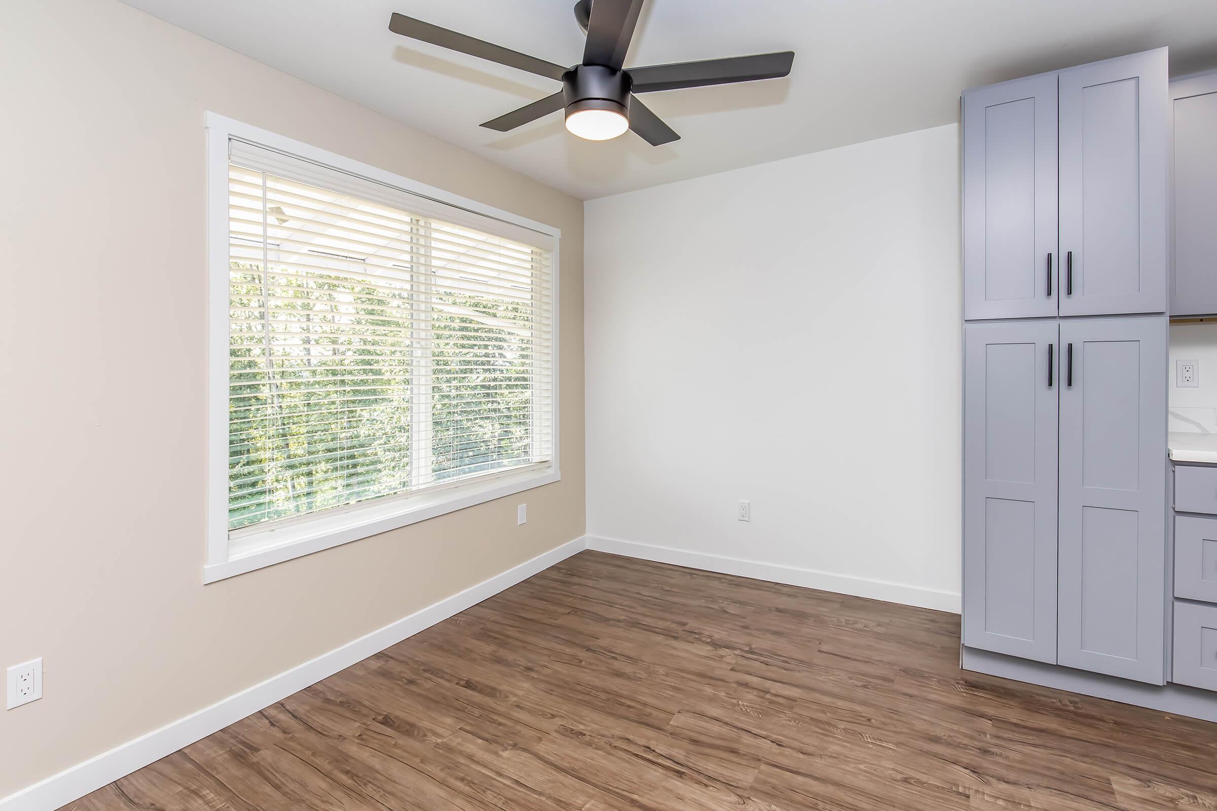 A bright, empty room with a large window featuring white blinds, a ceiling fan, and light wood flooring. One wall has tall, gray cabinets with a modern design. The space is clean and minimalistic, perfect for various uses or decoration.