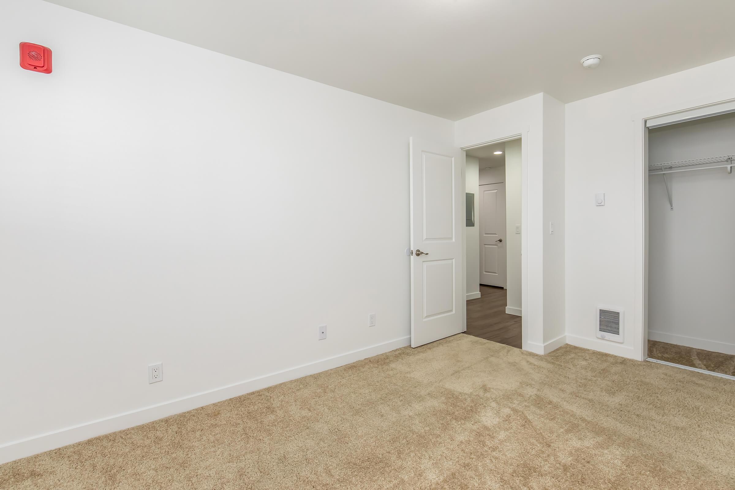 Empty room with light-colored walls and carpeted floor, featuring a closed door on the right leading to a hallway and a closet on the left. A smoke detector is visible on the far wall. The space is well-lit and minimalistic, suitable for furnishing.