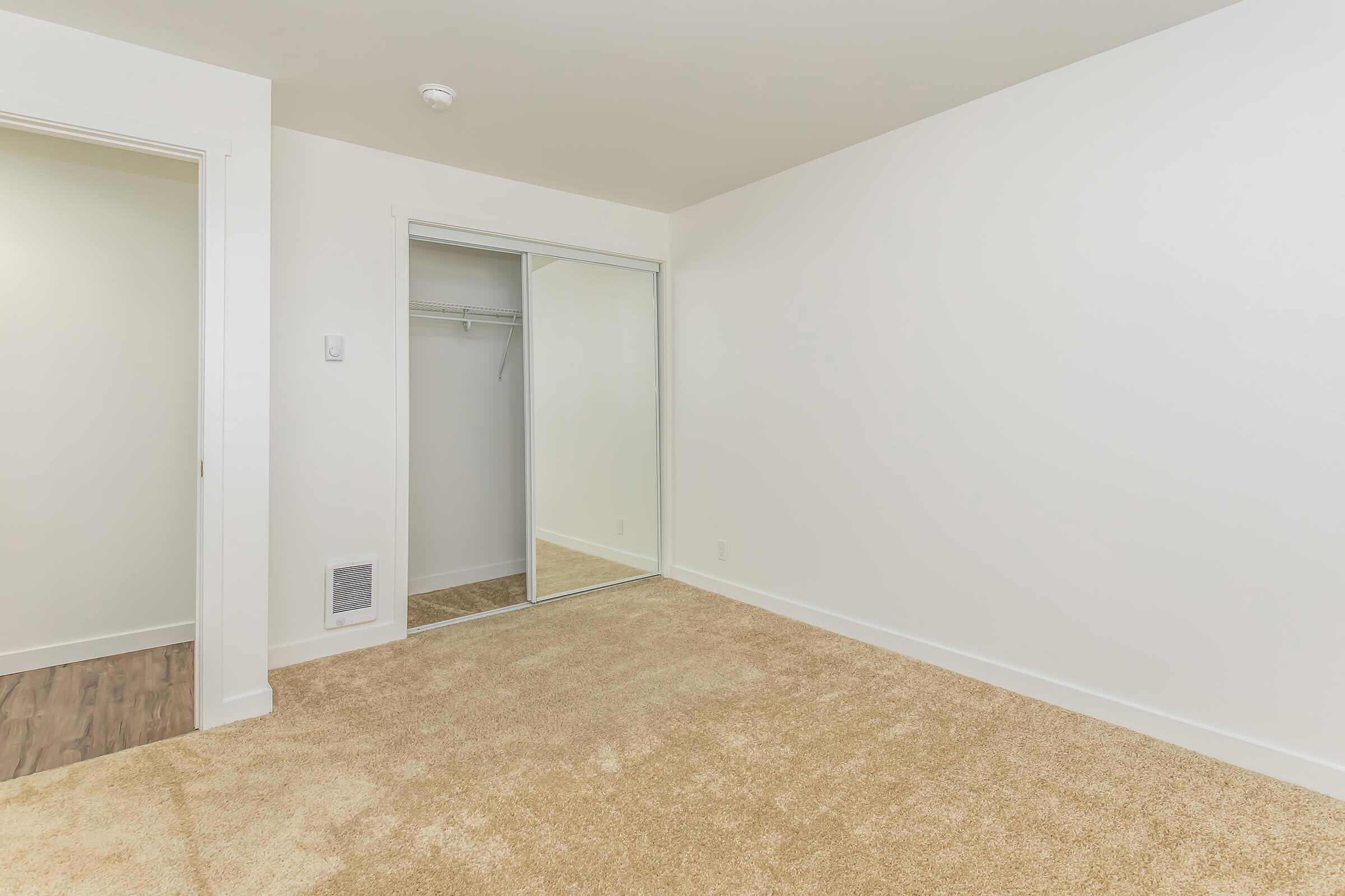 A spacious, empty room with light-colored walls and plush beige carpet. On one side, there's a sliding mirror closet door reflecting the room. The room features a doorway leading to an adjacent space, and a vent on the wall, creating a clean and uncluttered environment.