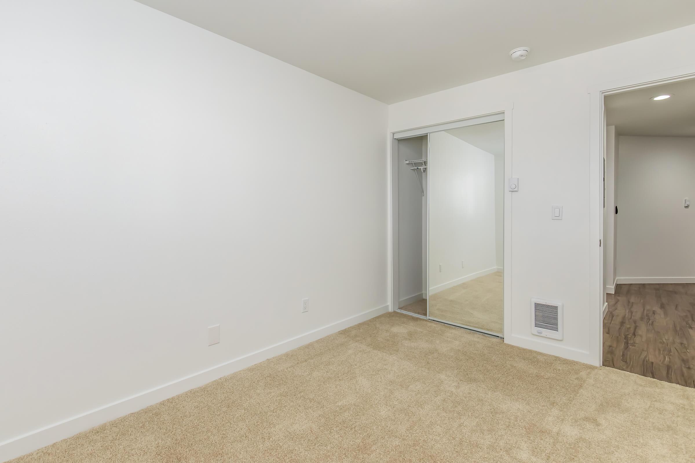 Empty room with beige carpet and white walls. A closet with mirrored doors is on the left side, and a door to another space is visible on the right. The room has a bright and neutral aesthetic, featuring an air vent on the wall. The overall atmosphere is calm and spacious.