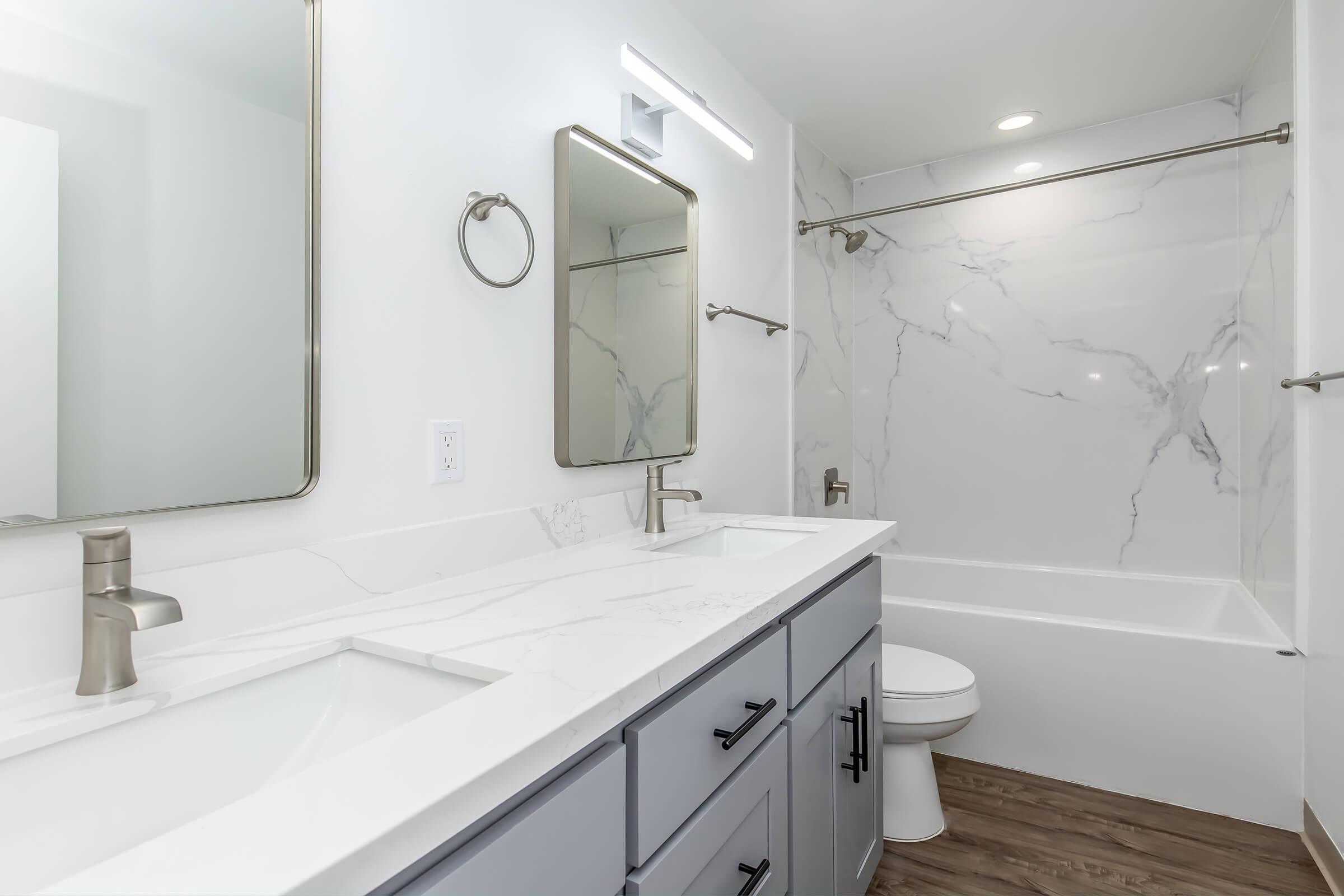 A modern bathroom featuring a double sink with a sleek countertop, a large mirror, decorative wall fixtures, and a bathtub with a marble-patterned surround. The decor includes light-colored walls, wooden flooring, and elegant lighting, creating a clean and sophisticated atmosphere.