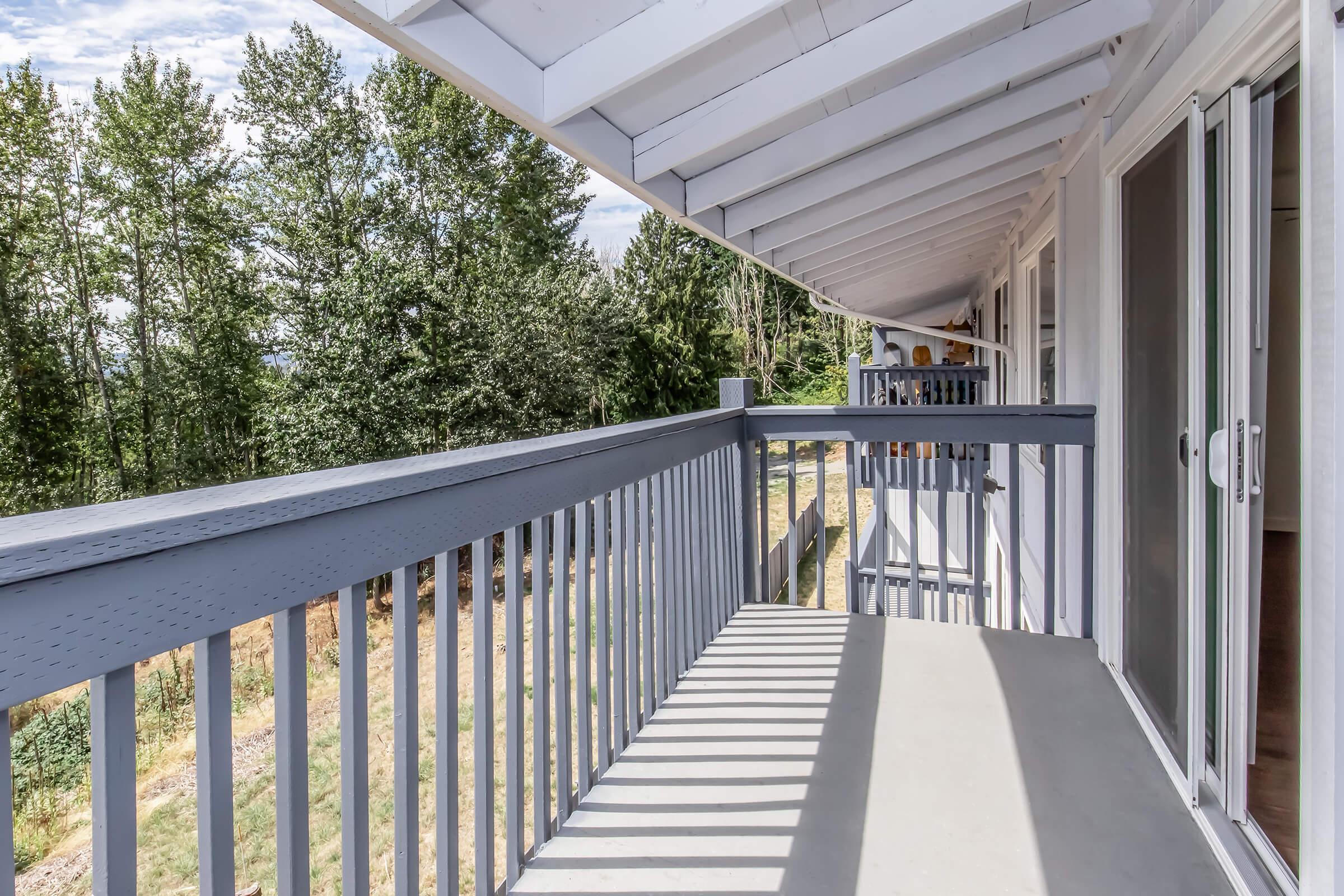 A balcony with a gray railing, featuring a concrete floor and shadows cast by overhead beams. In the background, there are lush green trees and a clear sky. The scene suggests a tranquil outdoor space, perfect for relaxation.