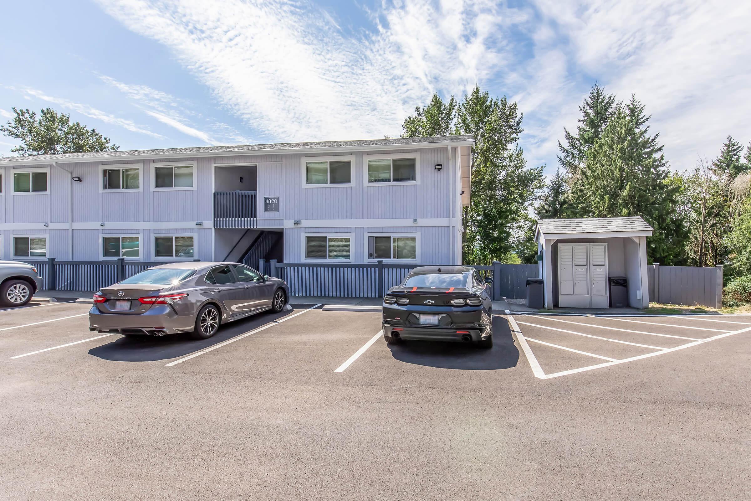 Parking area of a multi-story residential building with two cars parked in front. The building features multiple windows and a staircase leading to the upper level. Surrounding greenery and a storage shed are visible in the background under a clear blue sky.