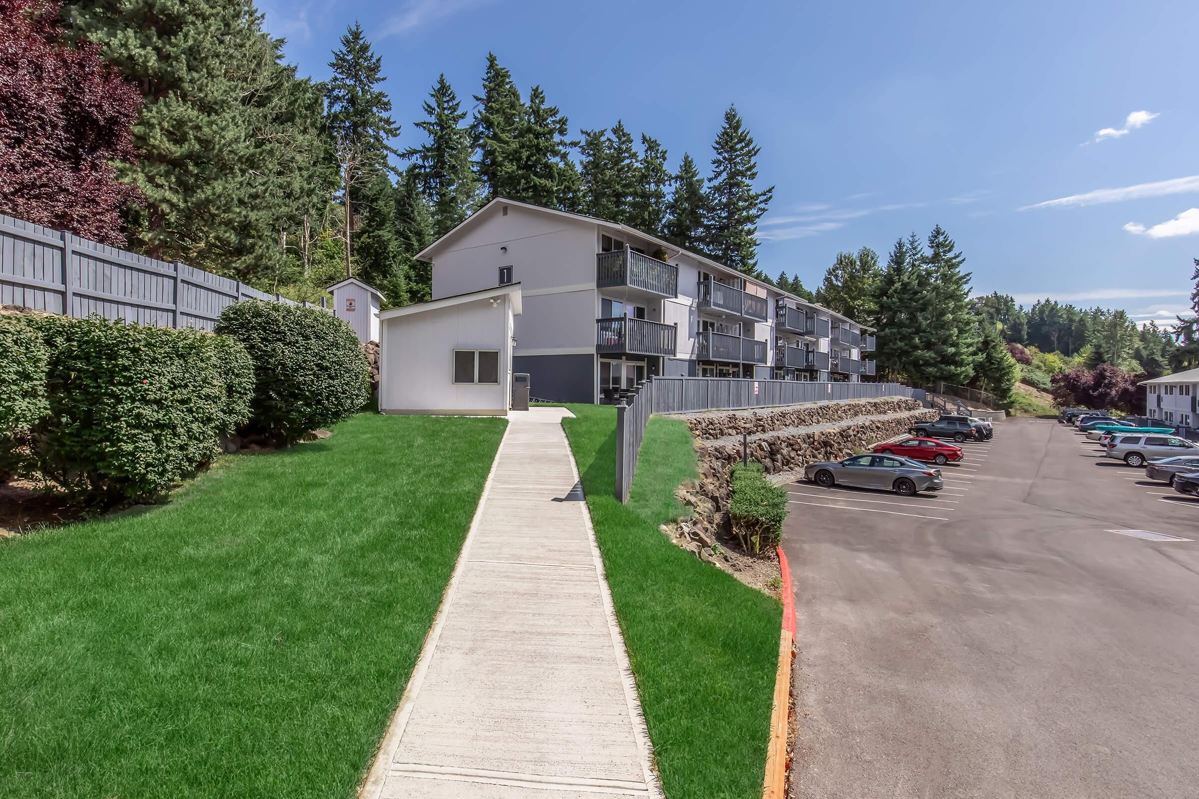 A modern apartment complex surrounded by greenery, featuring a well-maintained lawn and paved walkway. The building has multiple balconies and is set against a backdrop of tall trees. A parking lot is visible, with several cars parked. Bright blue sky with a few clouds adds to the pleasant atmosphere.
