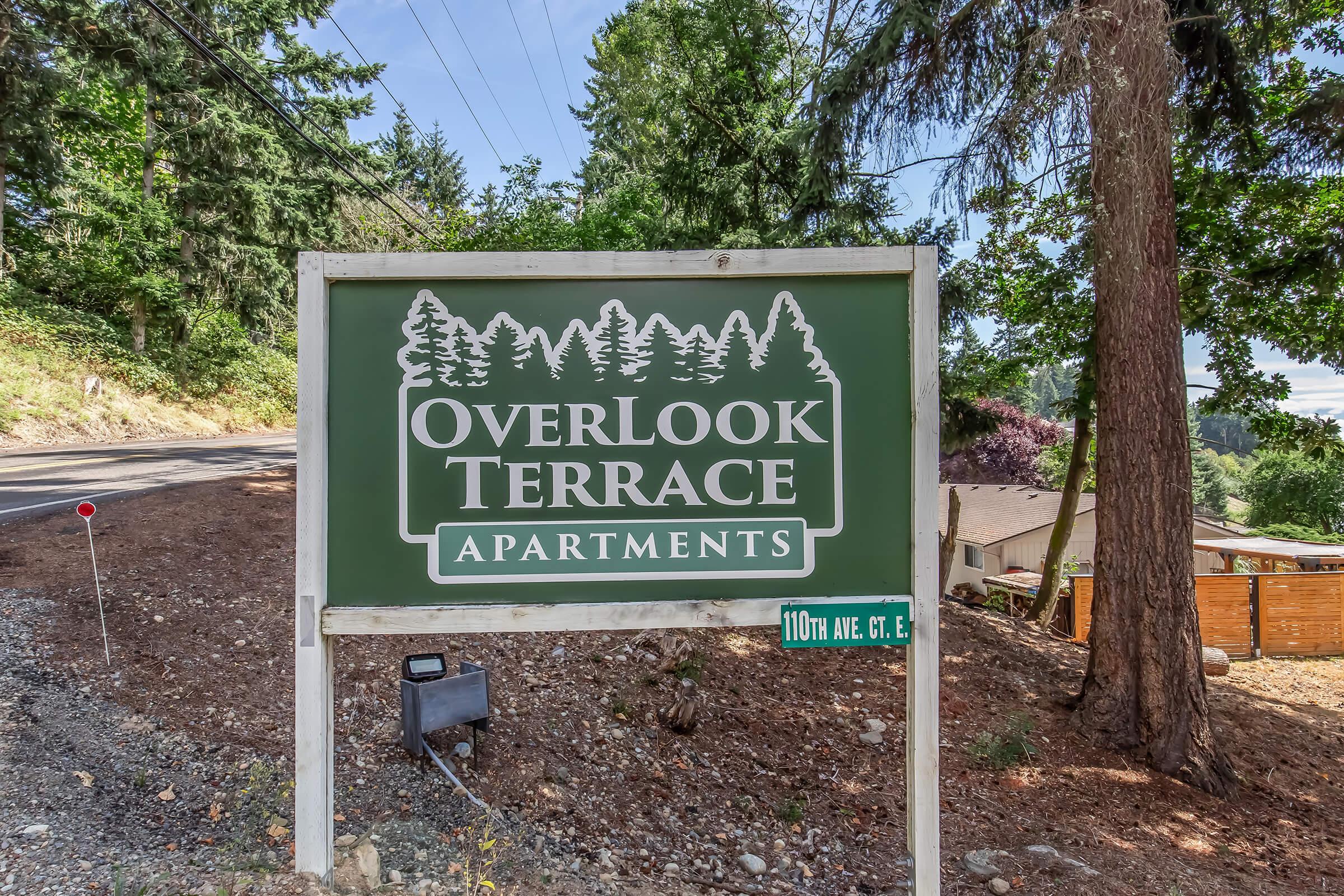 Sign for OverLook Terrace Apartments located at 110th Ave Ct E, surrounded by trees and a gravel road. The sign features a green background with white text and trees graphic, indicating the name and location of the apartments amidst a natural, wooded setting.