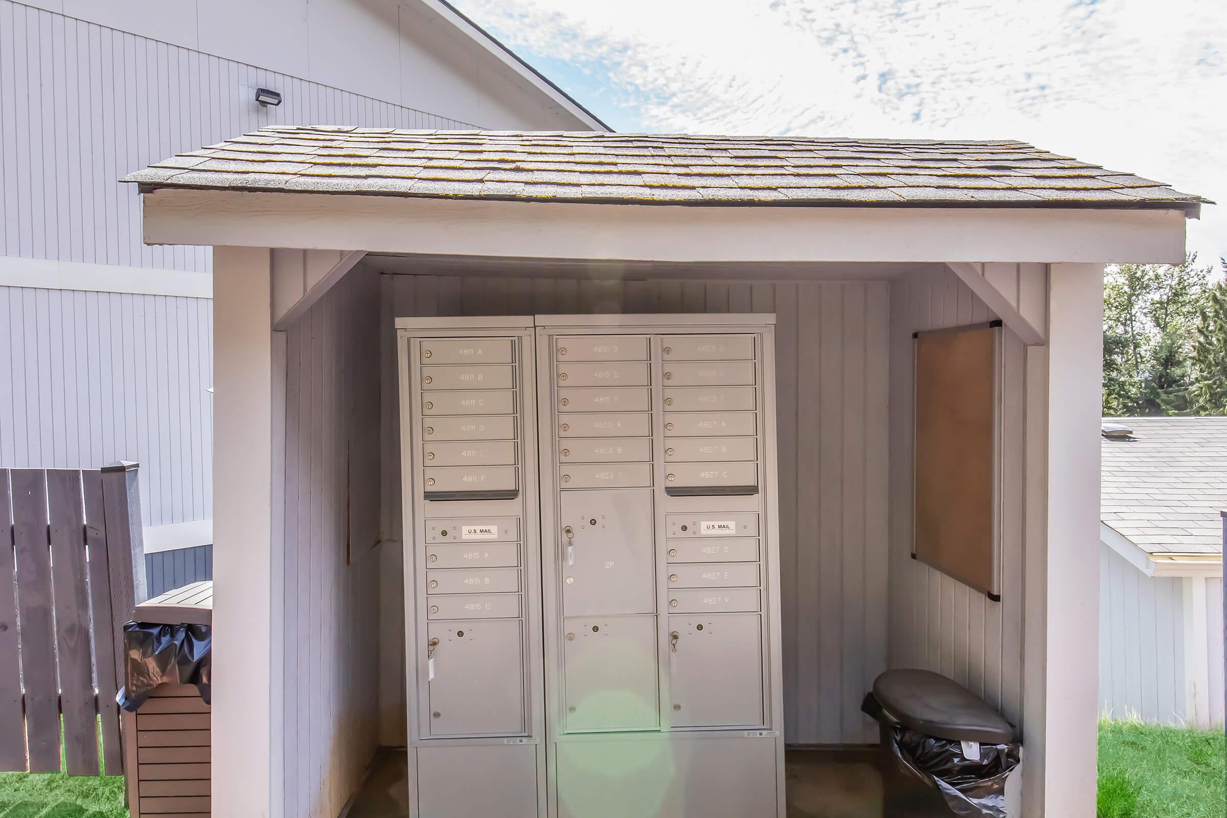 A weather-resistant mail kiosk with multiple secure mailboxes under a roof, positioned in a grassy area. A bulletin board is mounted on the side, and a trash can is visible nearby. The structure is framed by a light-colored building in the background and the sky is partly cloudy.
