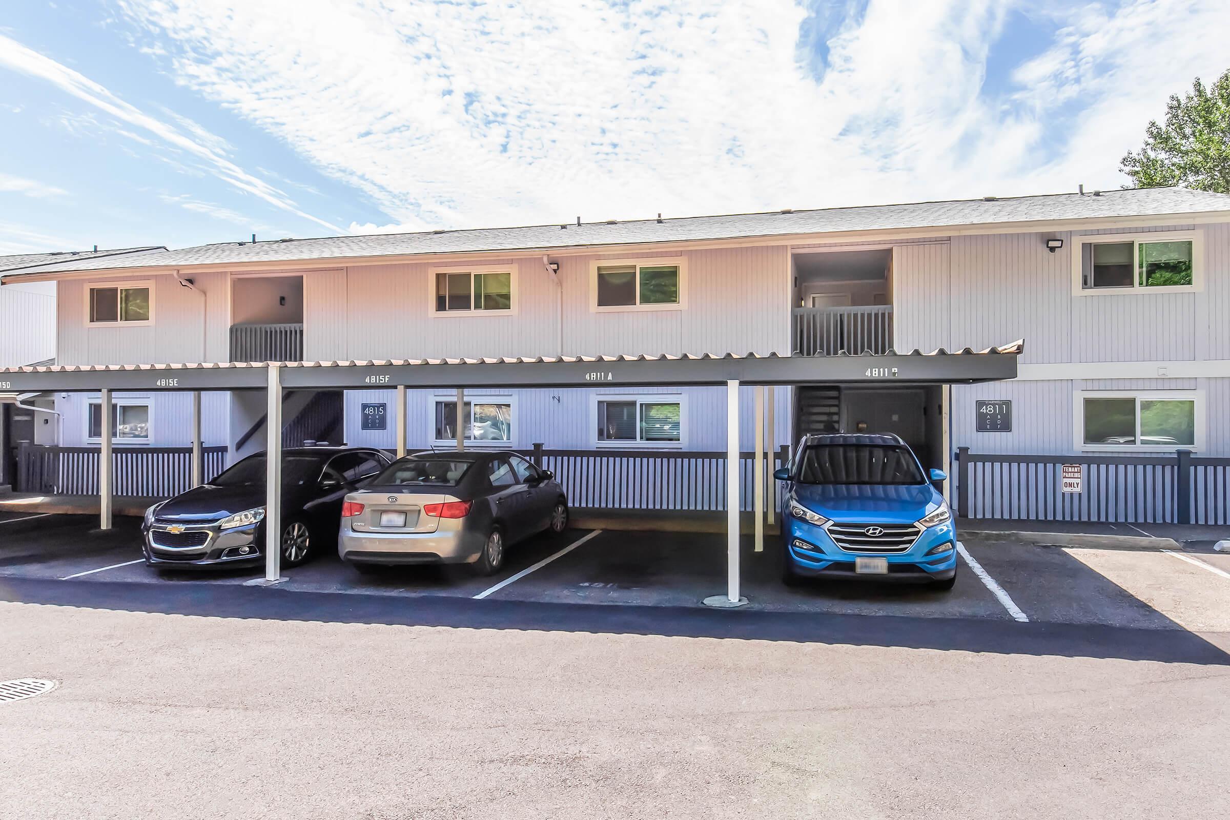 Two-story building with multiple apartment units featuring a white exterior and blue accents. Four parked cars are visible in front, with a covered area for two of them. The sky is clear with light clouds, and the surrounding area appears well-maintained.