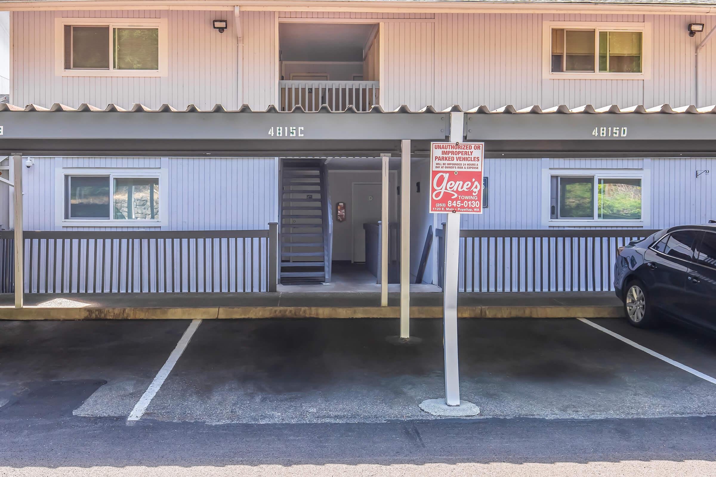 A multi-unit building with a covered porch area featuring staircases and windows. A parking lot is visible in the foreground, with one car parked in front of the building. A sign labeled "Gene's" is displayed prominently near the entrance, providing contact information.