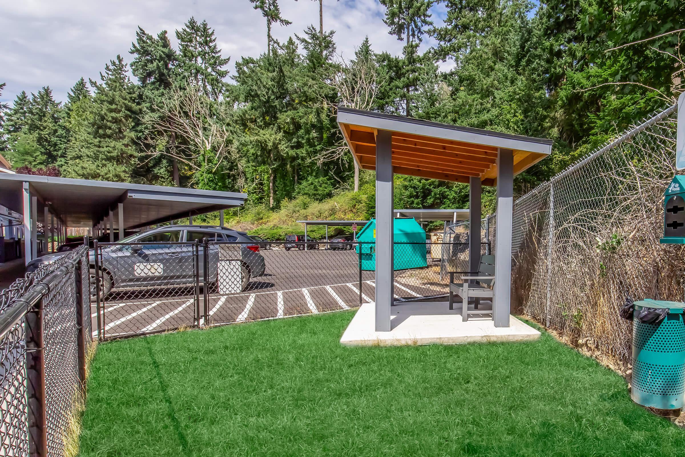 A view of a pet-friendly area featuring a small gazebo with a bench, surrounded by artificial grass. In the background, there are covered parking spaces and trees. A nearby waste station is visible, indicating amenities for pet owners. The setting is bright and inviting, designed for both pets and their owners.