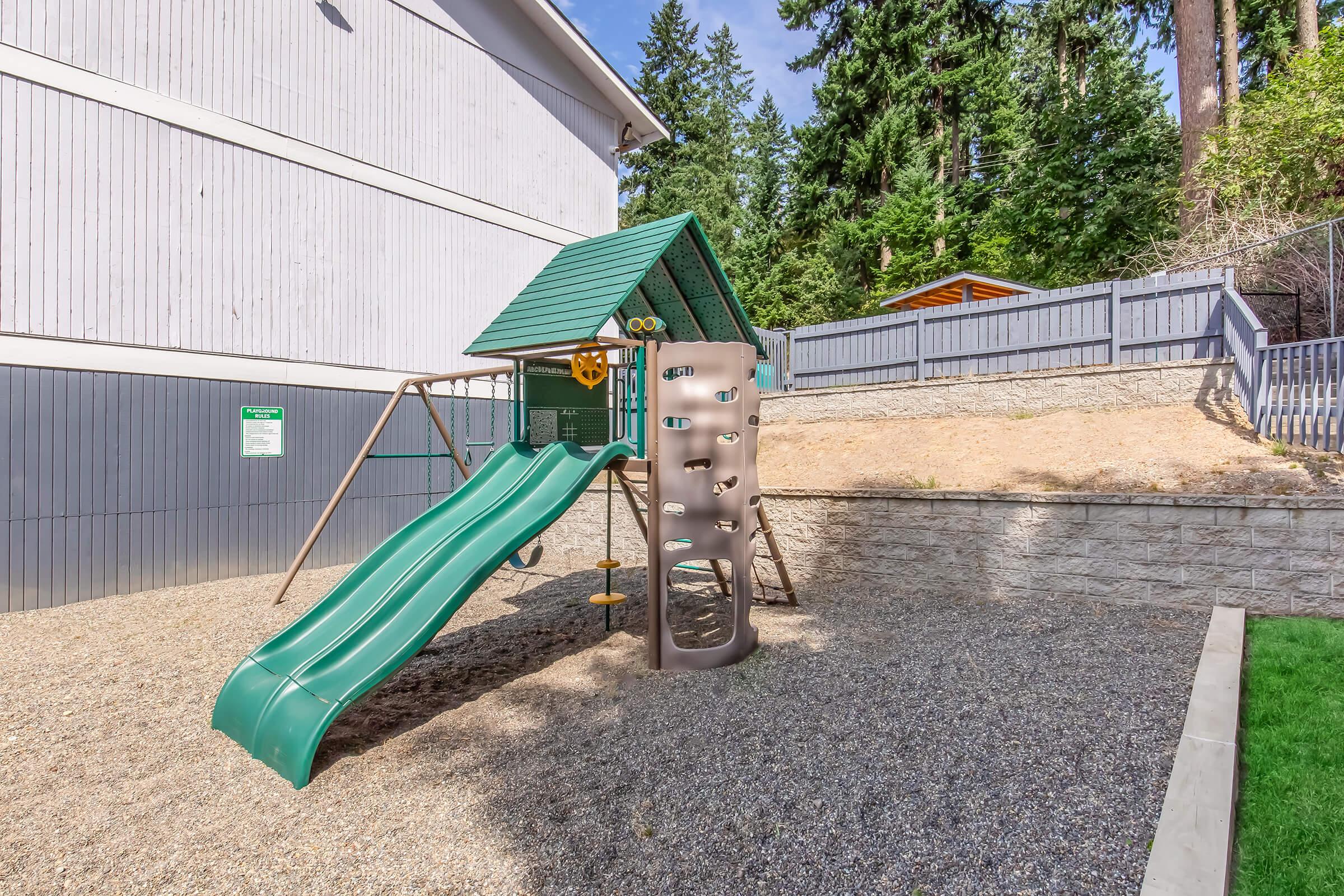 Playground equipment with a green slide and climbing structure, set on a gravel surface. It is located next to a building and surrounded by trees and a wooden fence. The area appears designed for children’s play.