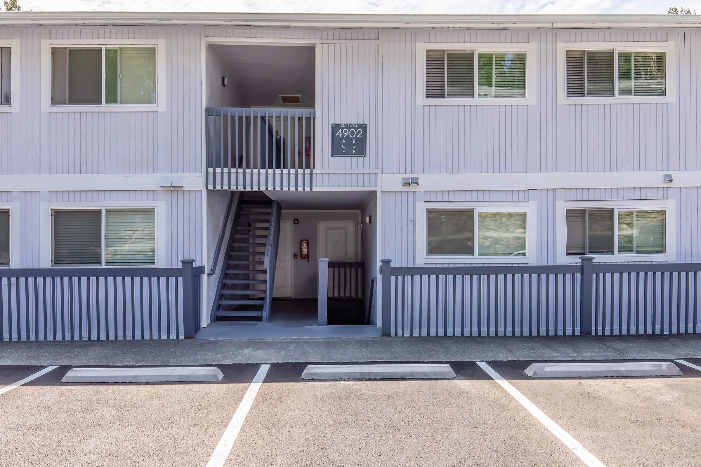 A two-story building with light gray exterior. The entrance is marked by the number "4902" and features a staircase leading to the second floor. Parking spaces are visible in front, with one space partially occupied. The sky is clear with a few clouds.