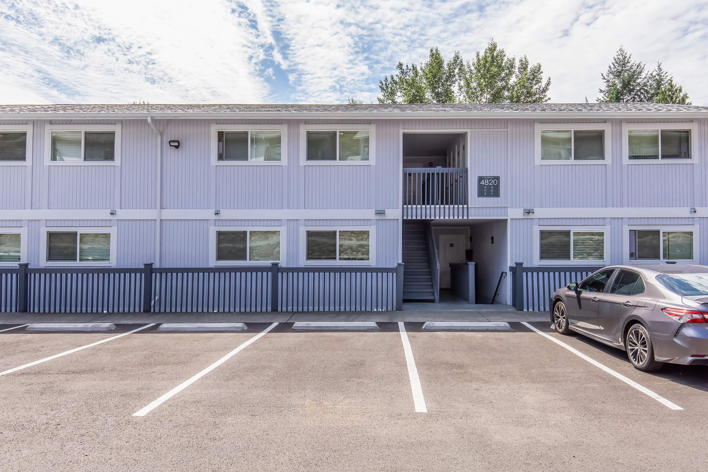 Exterior view of a two-story building with light purple siding. The building features several windows and a staircase leading to the upper floor. A silver sedan is parked in front, and there are marked parking spots visible. The sky is partly cloudy with some greenery in the background.