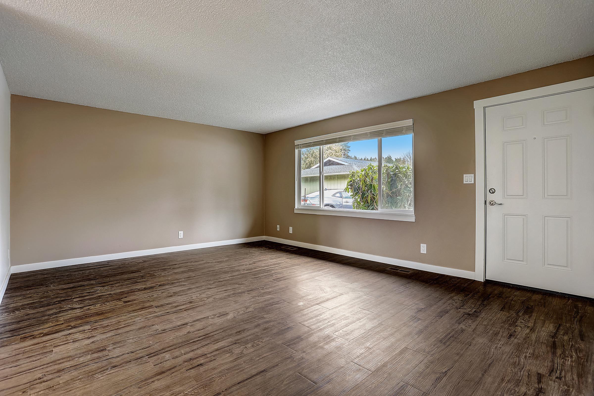 Empty room with brown walls and dark wooden flooring. A large window on the far wall lets in natural light, framing a view of greenery outside. A white door is visible on the right. The space is uncluttered, suggesting it’s ready for new furnishings or decoration.