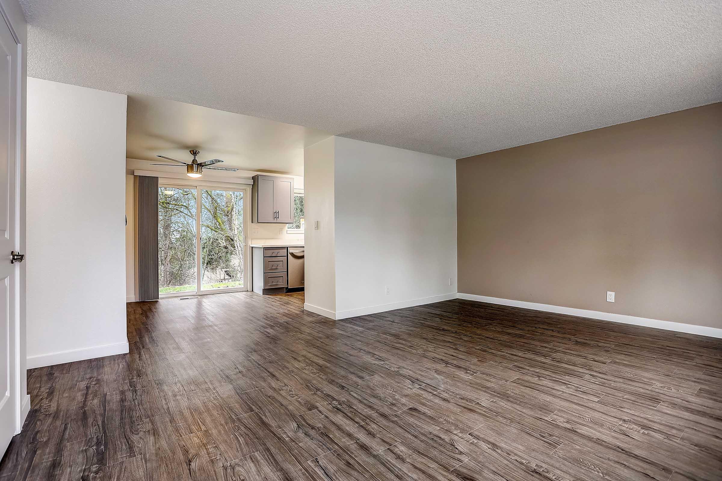 Spacious, empty living area with modern wood-like flooring and light-colored walls. To the left, a doorway leads to a kitchen with a ceiling fan, large window, and neutral cabinetry. Natural light fills the space, creating a bright and airy atmosphere.