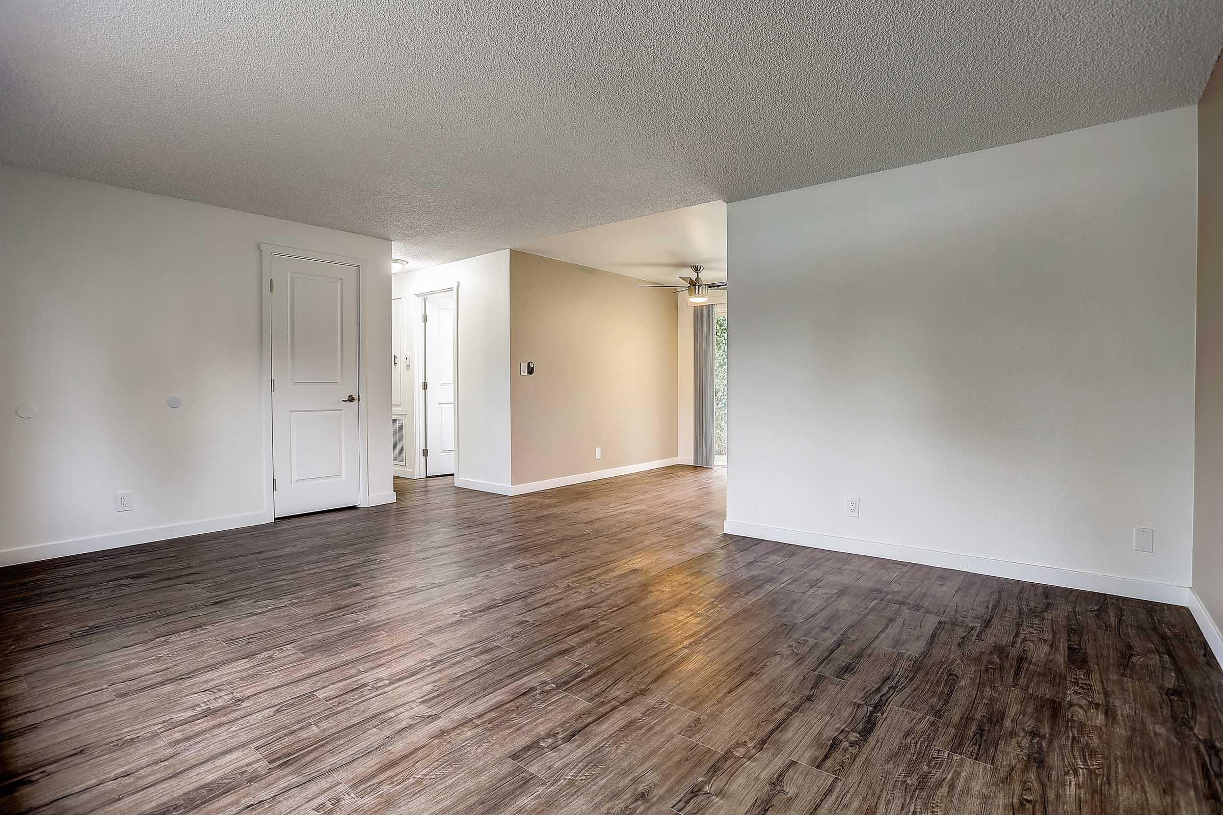 A spacious interior room with light-colored walls and a textured ceiling. The flooring is dark wood laminate. There is a closed white door on the left, an open doorway leading to another room in the back, and a ceiling fan visible in the distance. Natural light is coming from the right side.