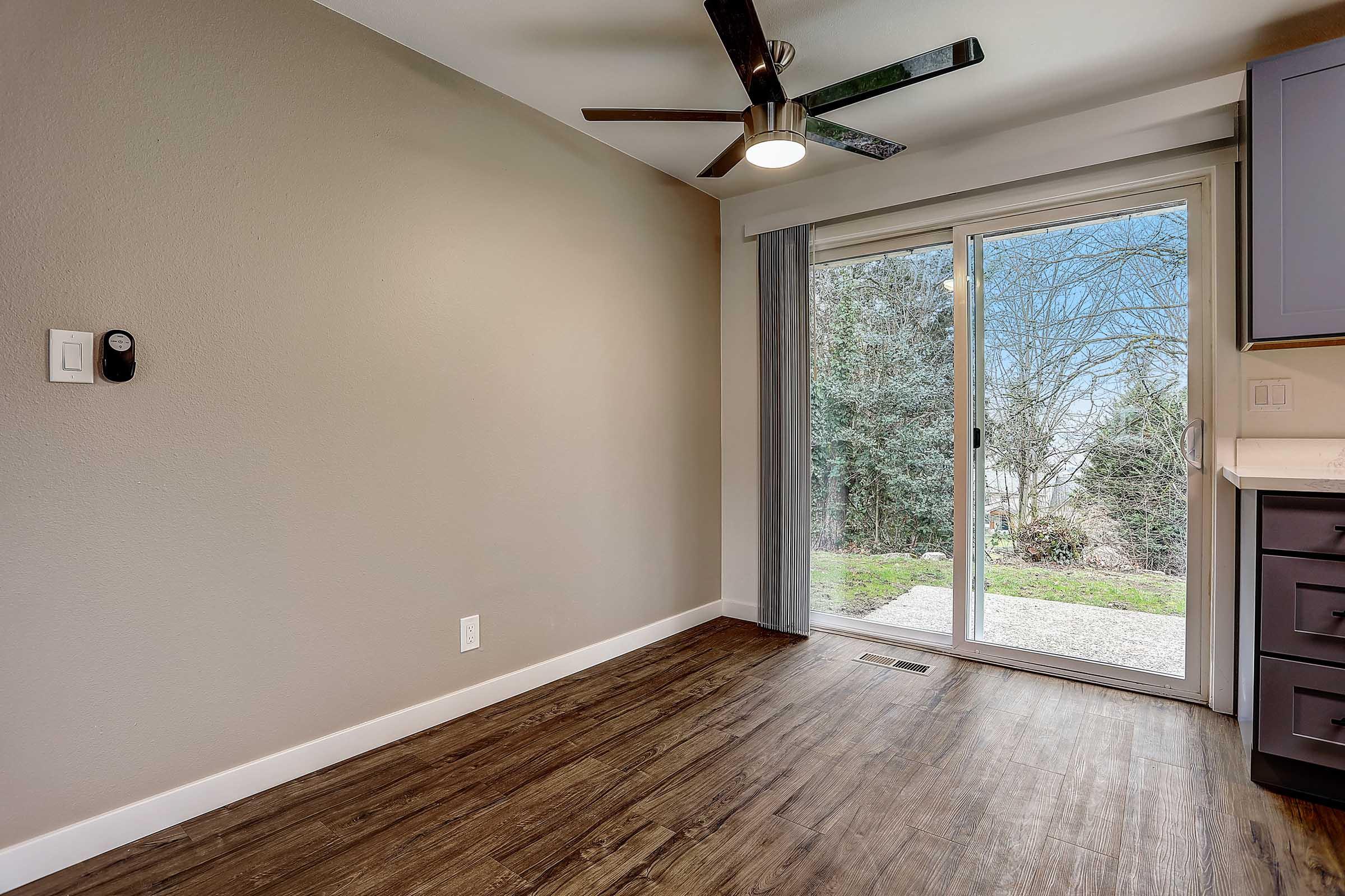 A modern, empty room featuring a ceiling fan, hardwood-style flooring, and neutral-colored walls. A sliding glass door leads to an outdoor area with trees visible outside. The space is well-lit with natural light and has a simple, contemporary design.