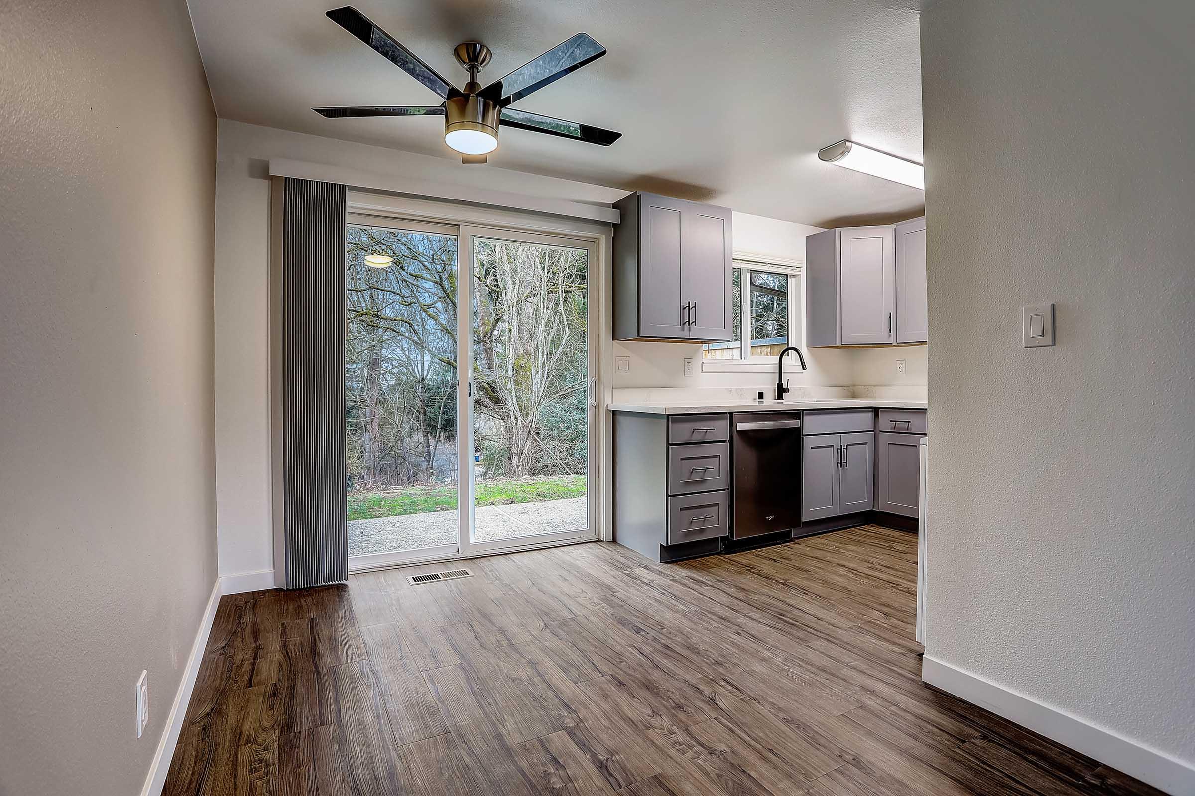 Interior view of a modern kitchen featuring gray cabinets, a stainless steel sink, and a dishwasher. A sliding glass door leads to a view of greenery outside. The kitchen has wooden flooring and overhead lighting, creating a bright and inviting space.