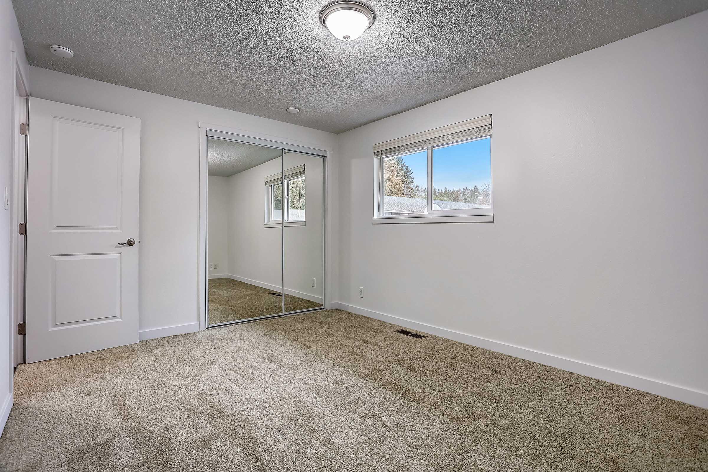 A clean, empty bedroom featuring light-colored walls, a large window with a view, and a mirrored closet. The floor is covered in plush carpet, and there is a ceiling light fixture. The overall atmosphere is bright and spacious.