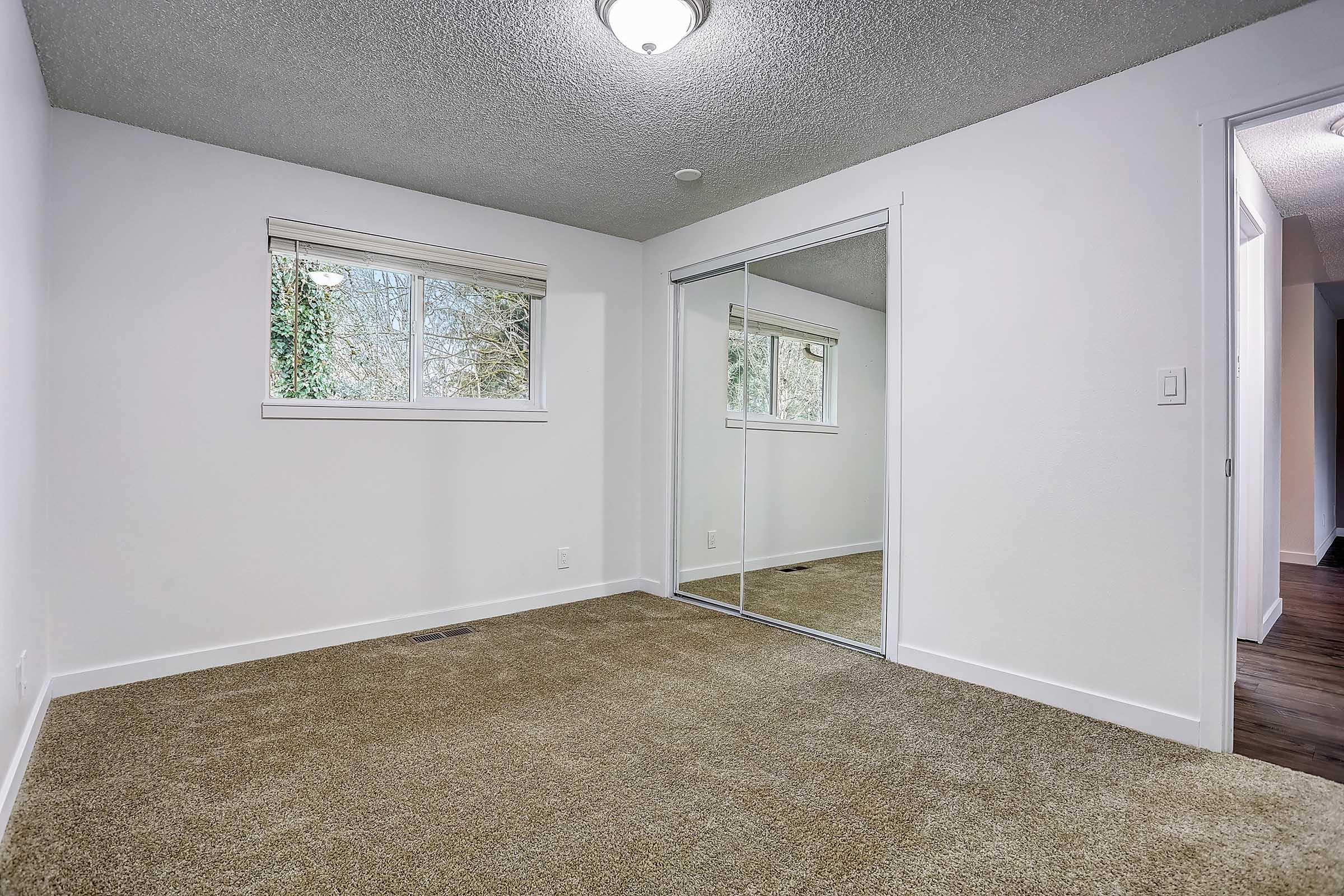 A well-lit, empty room featuring light-colored walls and a textured ceiling. There are two windows with natural light coming in, and a closet with mirrored doors. The floor is covered with beige carpet, giving a warm and inviting feel to the space.