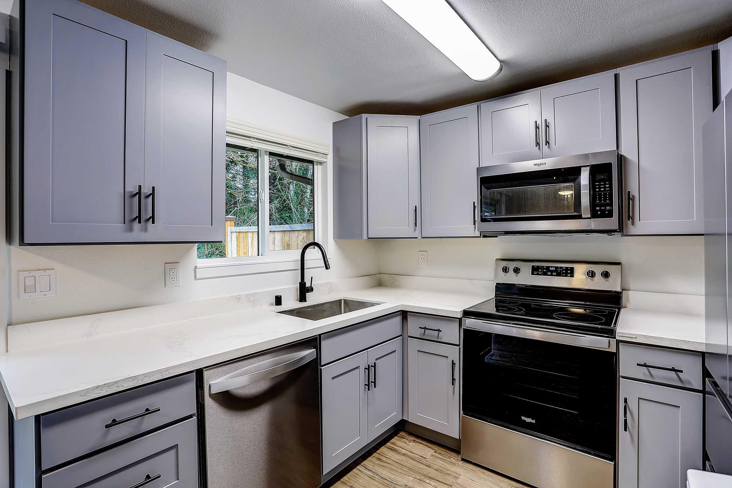 Modern kitchen with light gray cabinetry, stainless steel appliances, and white countertops. Features include a microwave, an electric stove, a sink with a black faucet, and a window allowing natural light. The flooring is a warm wood tone, creating a cozy atmosphere.