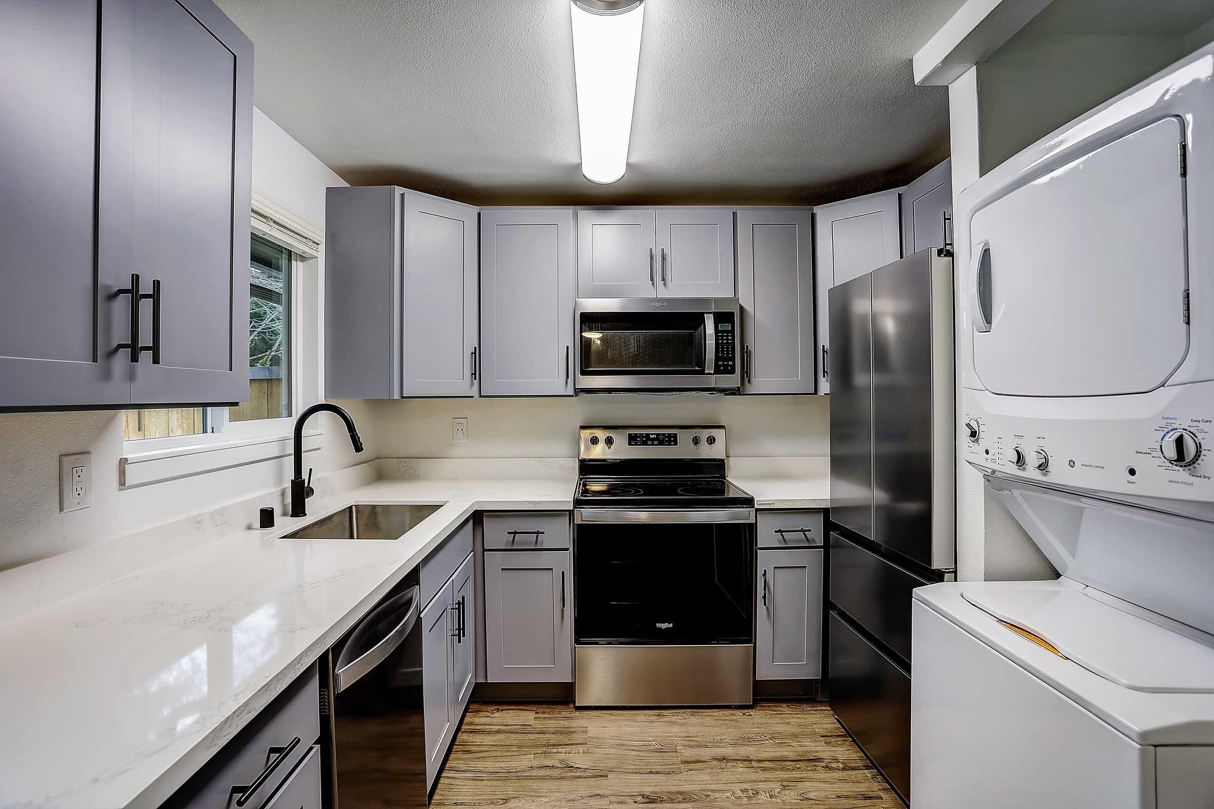 Modern kitchen featuring gray cabinets, a stainless steel microwave, oven, and refrigerator. The countertop is light-colored, and a sleek sink is installed. A compact washer and dryer are located to the right, with natural light coming from a window. Stylish and functional design elements are present throughout the space.
