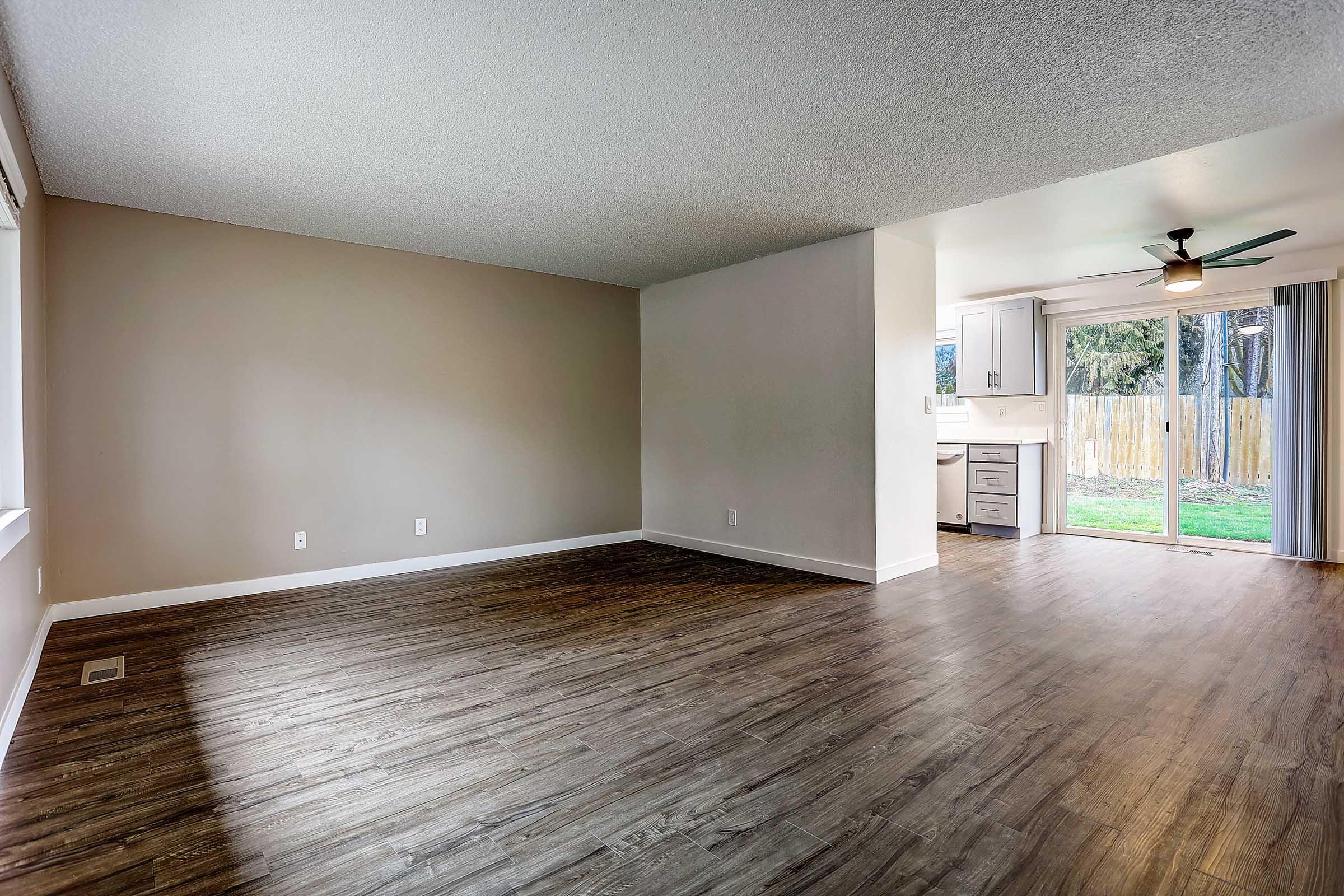 Spacious living room with hardwood-style flooring, large windows allowing natural light, and a neutral color palette. Features a ceiling fan and an open layout leading to a modern kitchen with white cabinetry and access to a backyard.