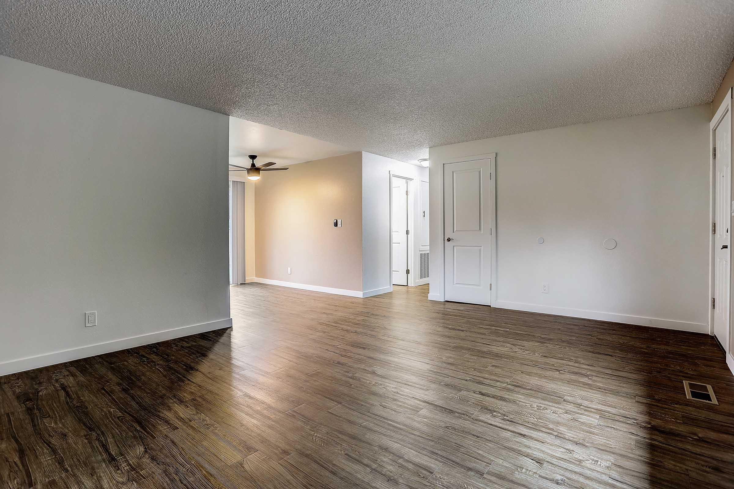 Spacious interior of an unfurnished room featuring wood-style flooring, a ceiling fan, and neutral-colored walls. A doorway leads to another room, with natural light entering from a window. The layout is open, providing a versatile space for various arrangements.