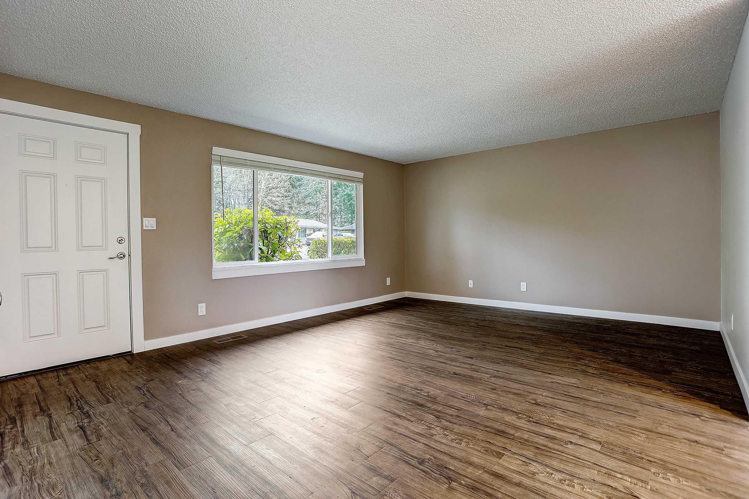 A spacious, empty living room featuring light brown walls, a large window letting in natural light, and a wooden floor. The door on the left leads outside, and there are no furnishings, creating an inviting and open atmosphere.