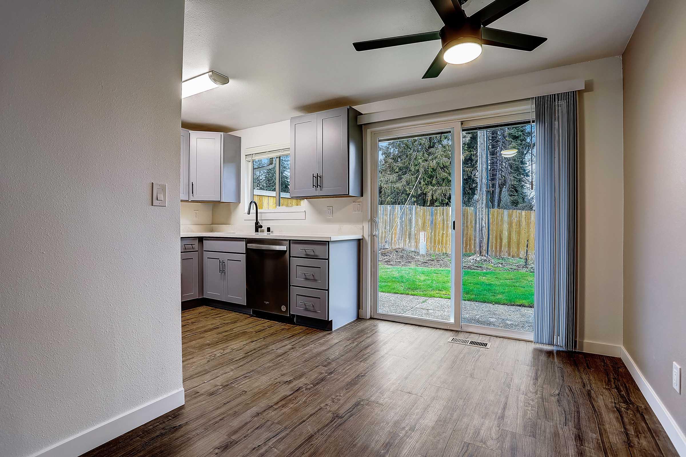 Modern kitchen with gray cabinets, stainless steel appliances, and a sliding glass door leading to an outdoor space. The room features a ceiling fan, hardwood-style flooring, and a bright atmosphere with natural light streaming in. The backyard is visible through the glass door, showcasing a grassy area.