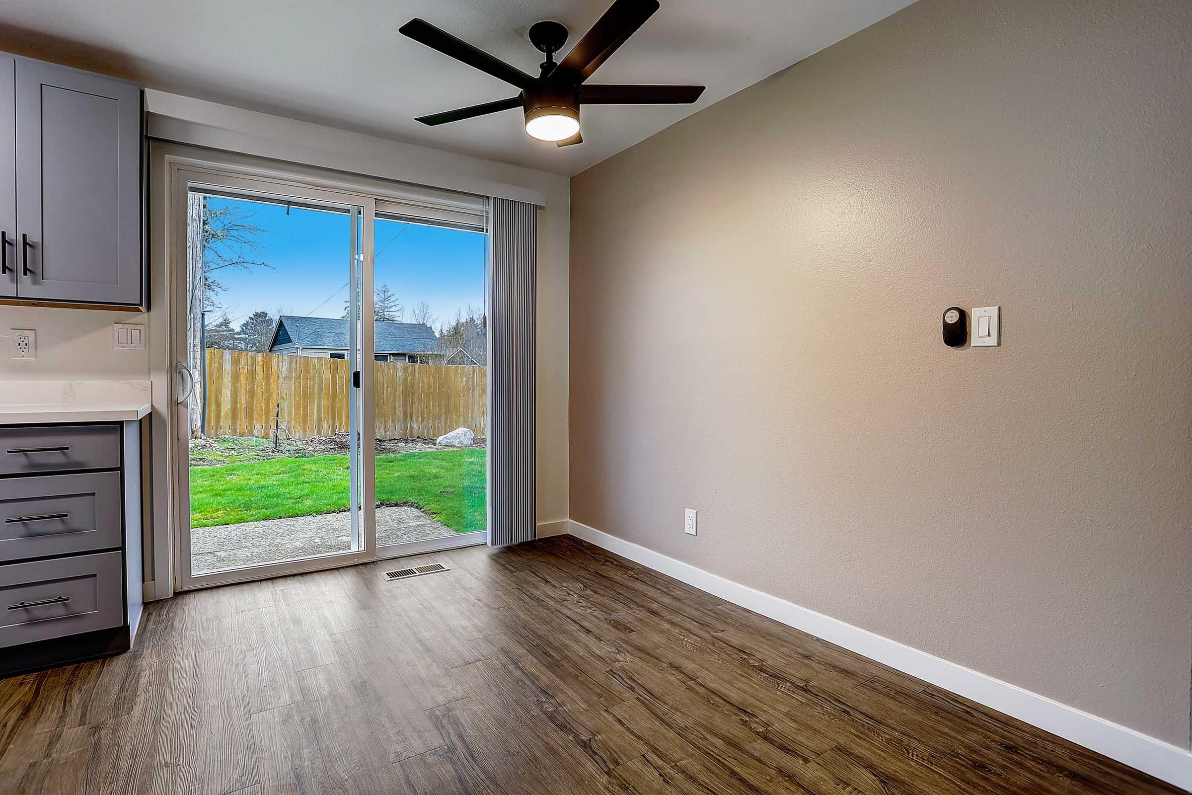 Interior view of a room featuring a ceiling fan, large sliding glass door with a view of an outdoor area, wood-like flooring, and modern kitchen cabinets. The walls are painted a soft gray, and natural light floods the space, creating a warm and inviting atmosphere.