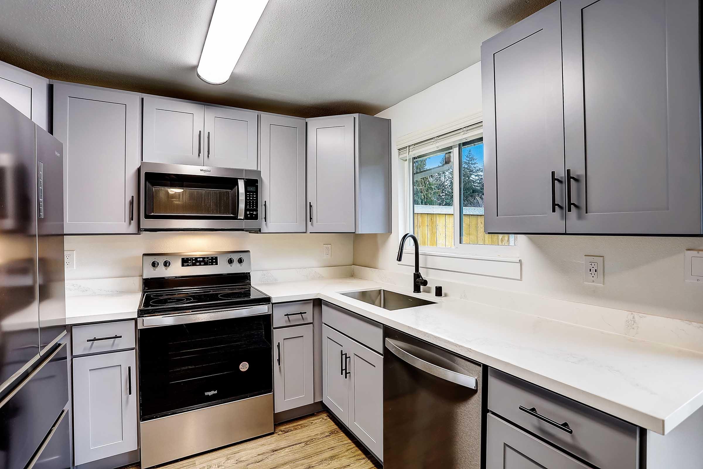 Modern kitchen featuring gray cabinets, stainless steel appliances, a black oven and microwave, a double sink, and a large window allowing natural light in. The countertops are white, complemented by wooden flooring, creating a sleek and contemporary look.