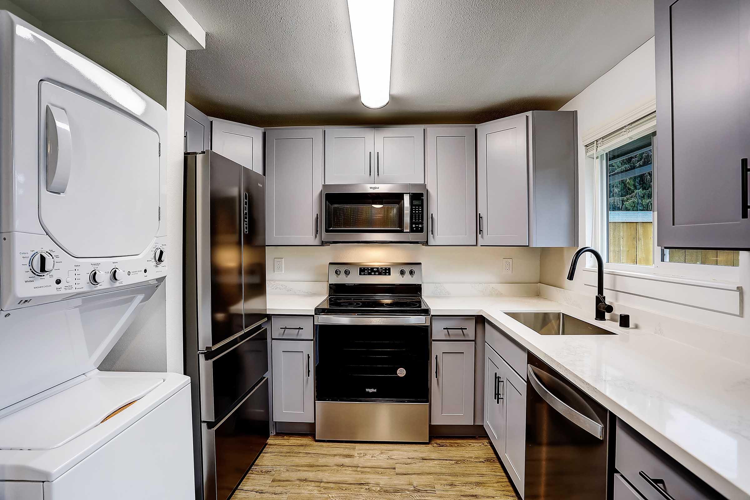 Modern kitchen featuring gray cabinets, stainless steel appliances including a refrigerator, microwave, and stove. A washer and dryer are visible on the left. The countertop is white with a subtle pattern, and there's a window providing natural light. The flooring is light wood, creating a warm atmosphere.