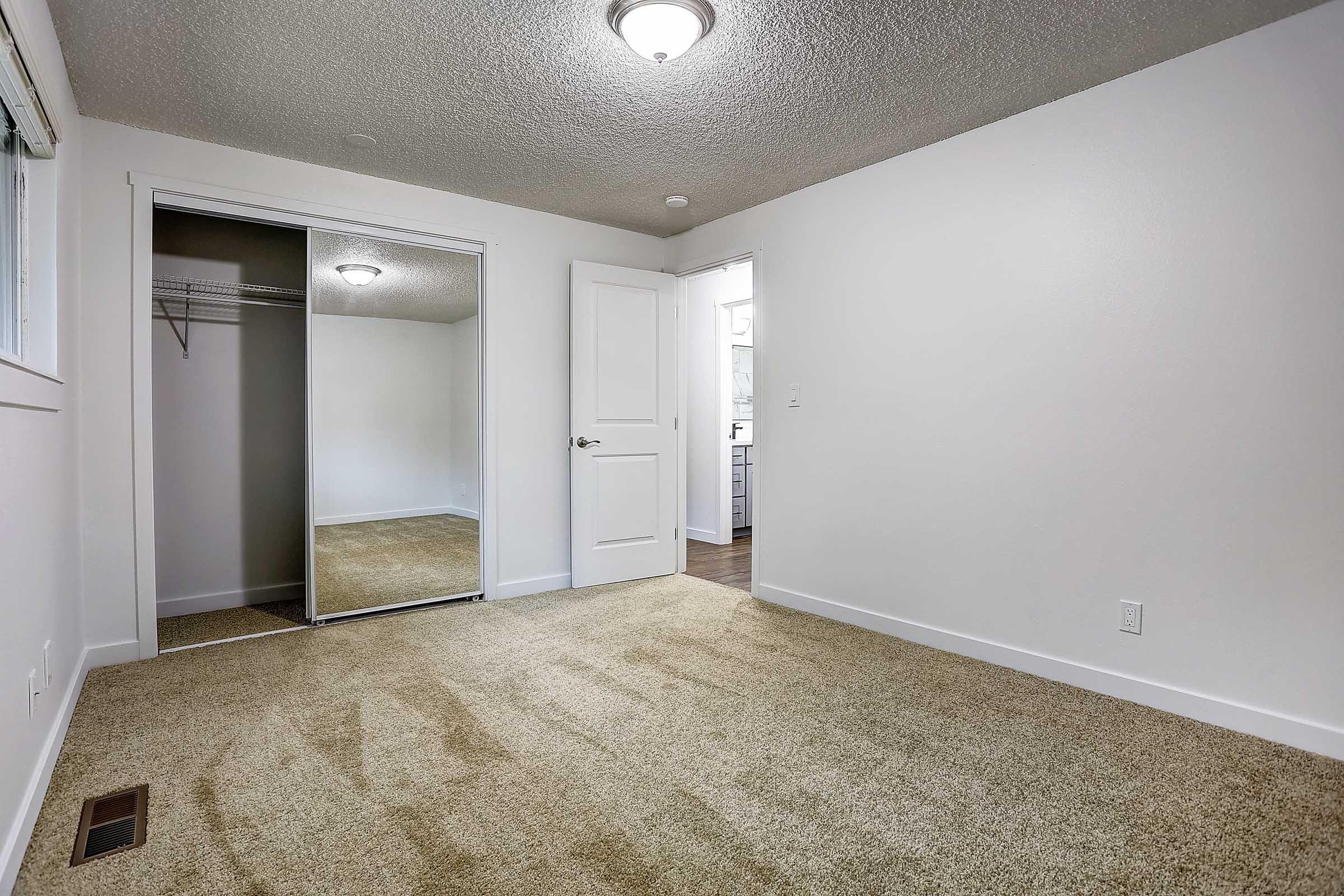 A spacious, empty room featuring beige carpet, a mirrored sliding closet door, and a ceiling light fixture. The walls are painted white, and there is a doorway leading to another room visible in the background. Natural light is coming from a window on the left side.