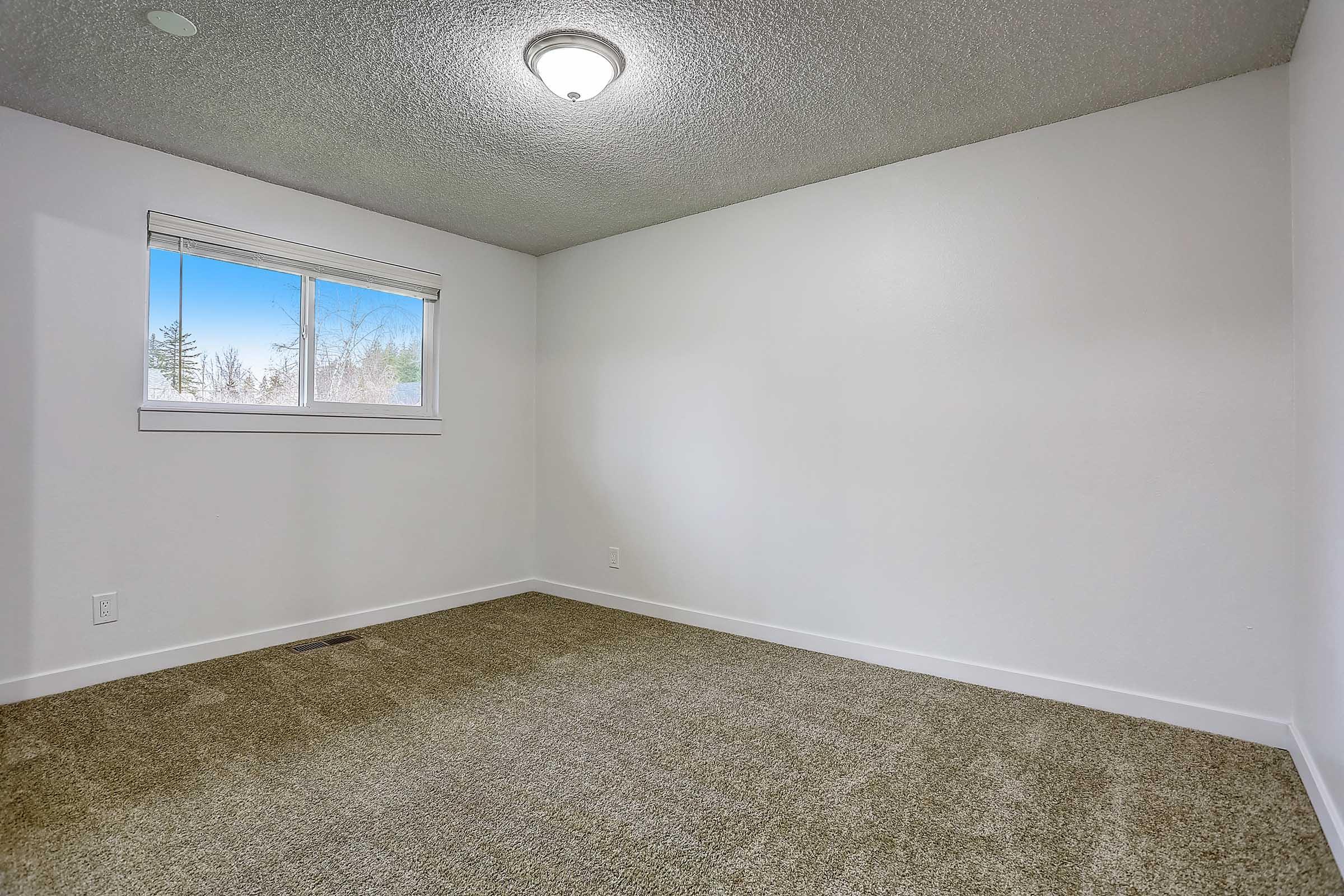 A spacious, empty room with light-colored walls and a textured ceiling. The floor is covered in carpet, and there is a single window on one wall, allowing natural light to enter. The room has a simple pendant light fixture in the center of the ceiling.
