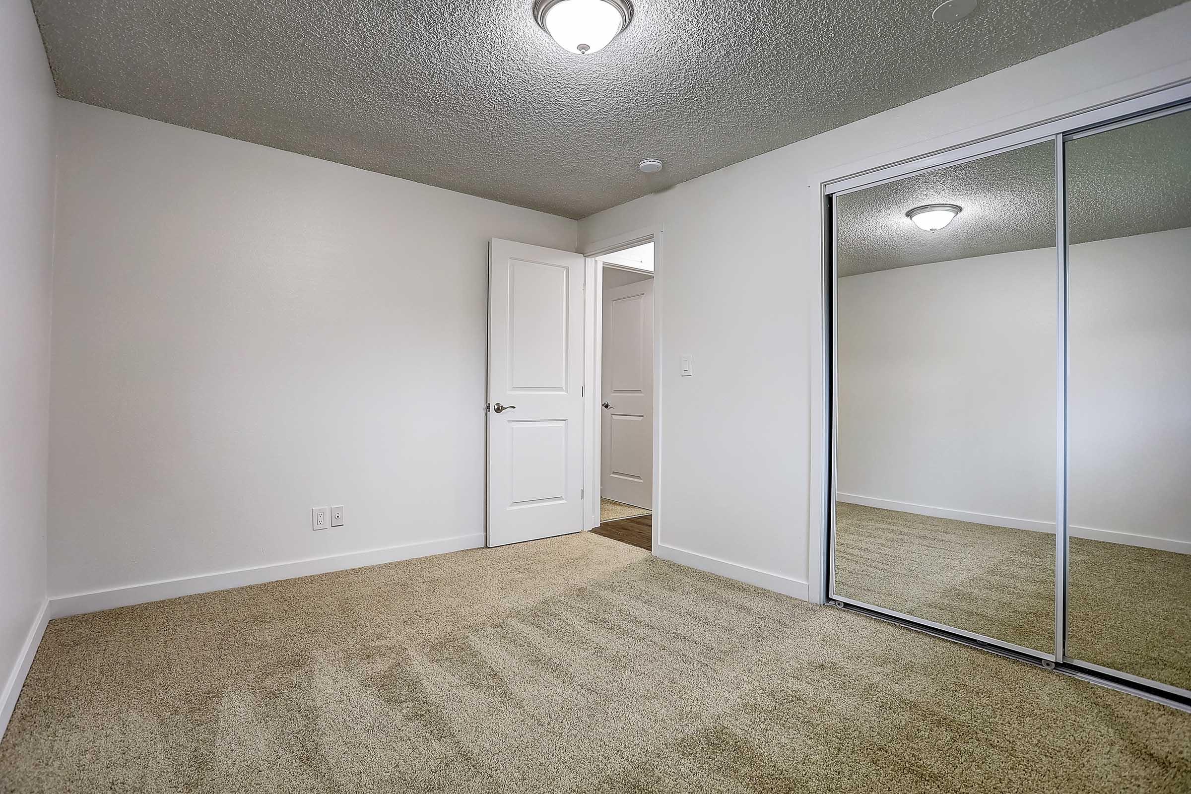 A vacant bedroom featuring beige carpet, a ceiling light fixture, and a white door leading to a hallway. There is a large mirrored closet against one wall, and the room has neutral-colored walls, creating a bright and spacious atmosphere. The door to the bathroom is partially visible in the background.