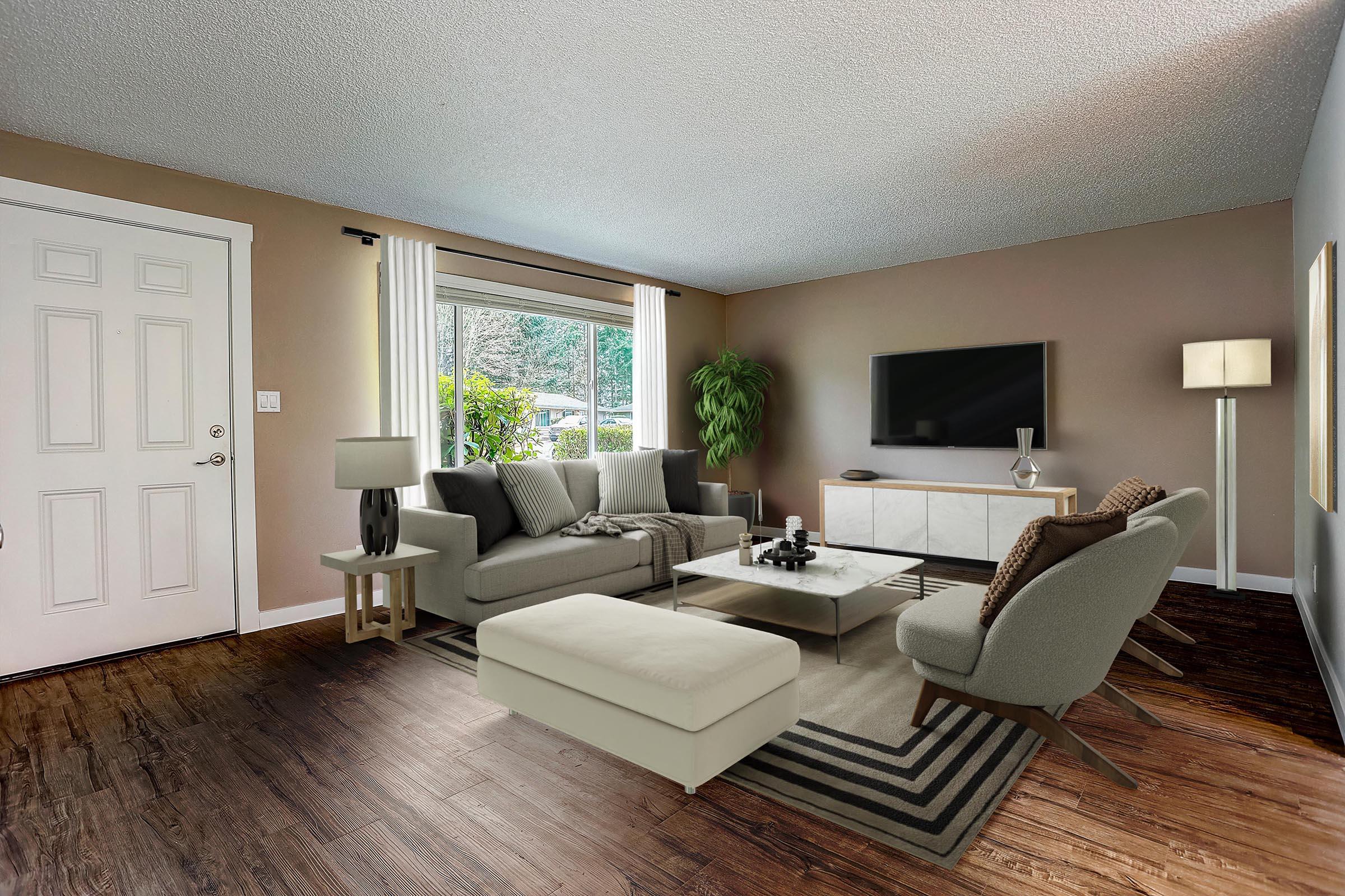 A modern living room featuring a light gray sofa, a neutral coffee table, and two stylish armchairs. A large window lets in natural light, revealing green outdoor foliage. There's a sleek TV mounted on the wall, a floor lamp, and a potted plant, all set against warm, earthy tones and wooden flooring.