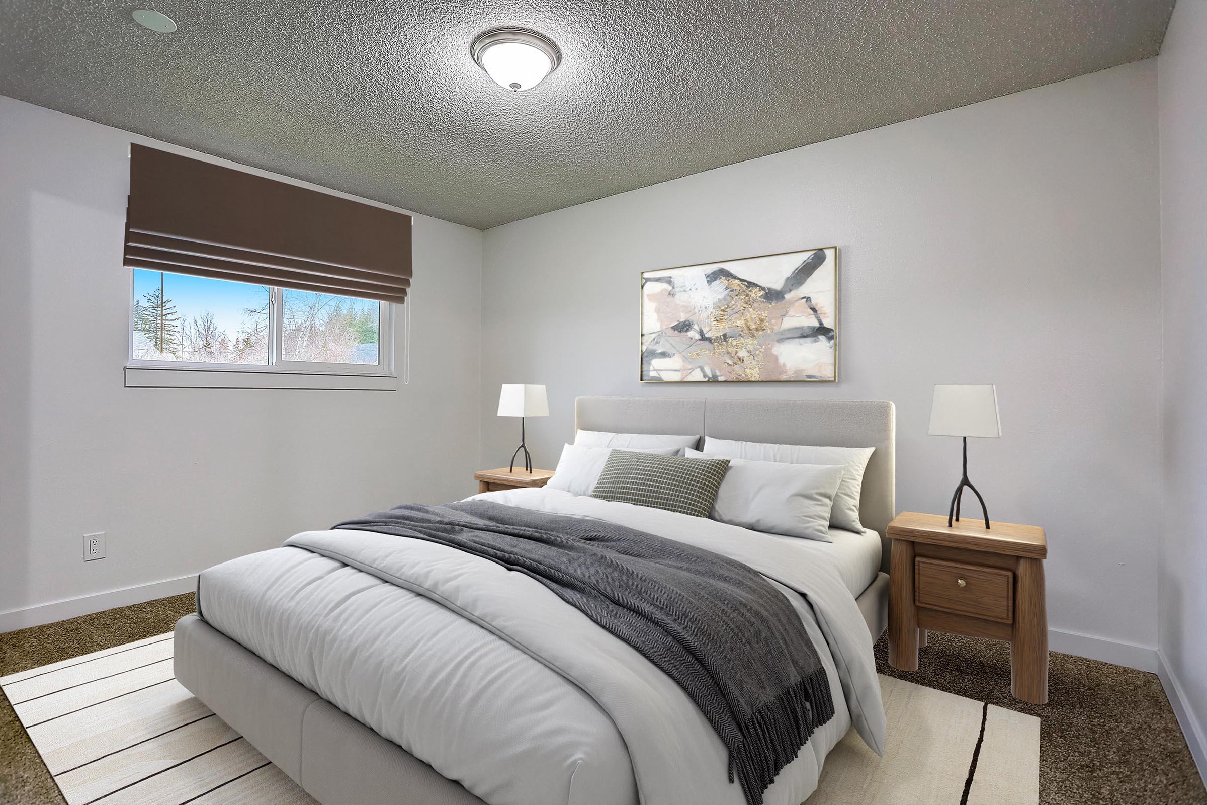 A cozy, modern bedroom featuring a queen-sized bed with white bedding and a gray throw blanket. There are two wooden nightstands with lamps on either side of the bed. A large window lets in natural light, and a piece of abstract art hangs on the wall, adding a touch of color to the neutral decor.