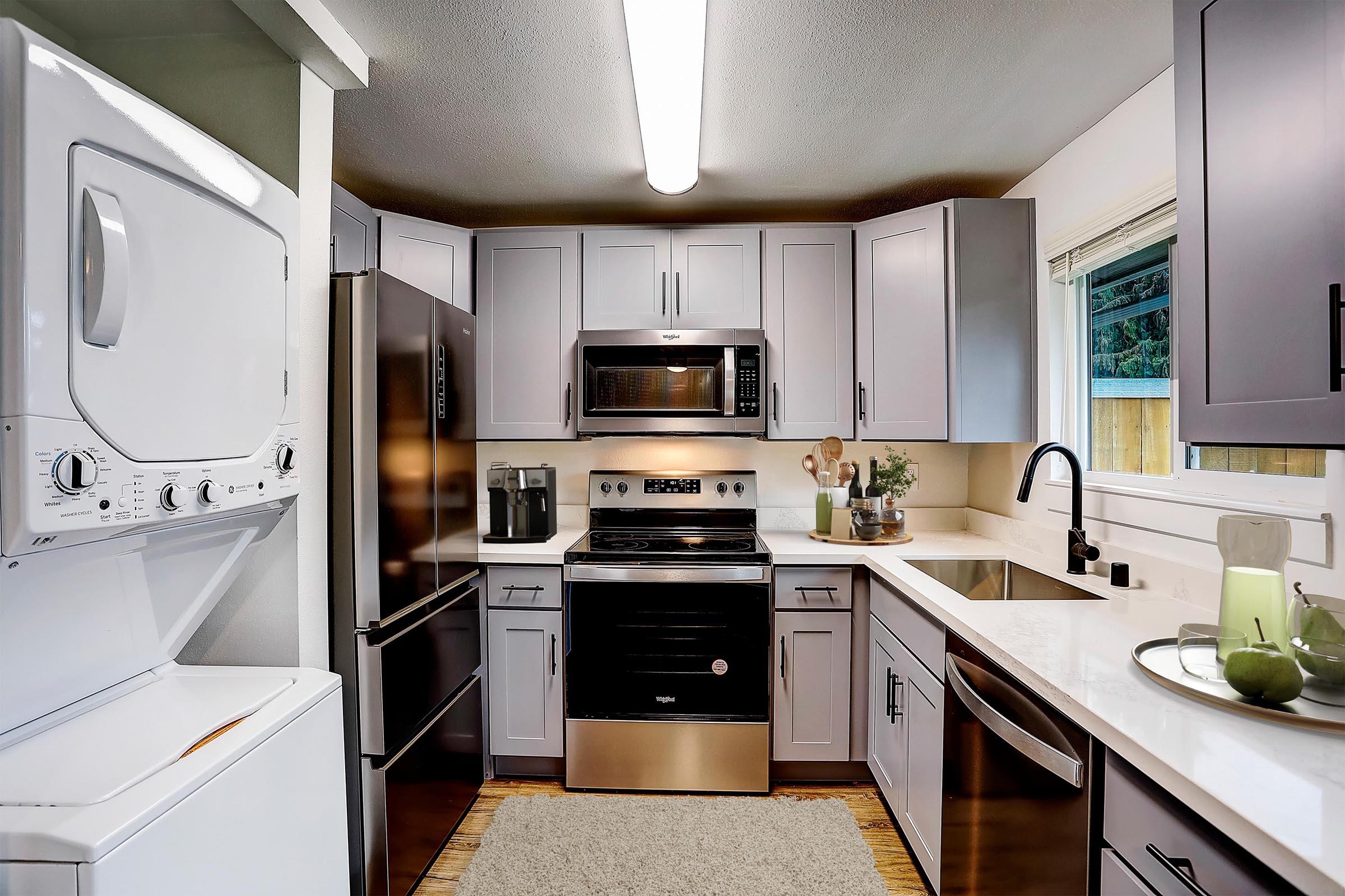 A modern kitchen featuring gray cabinets, stainless steel appliances including a refrigerator, oven, and microwave. There is a washing machine and dryer combo in the corner, a sink with a black faucet, and a window providing natural light. Neutral-colored decor and a countertop with kitchen accessories complete the space.