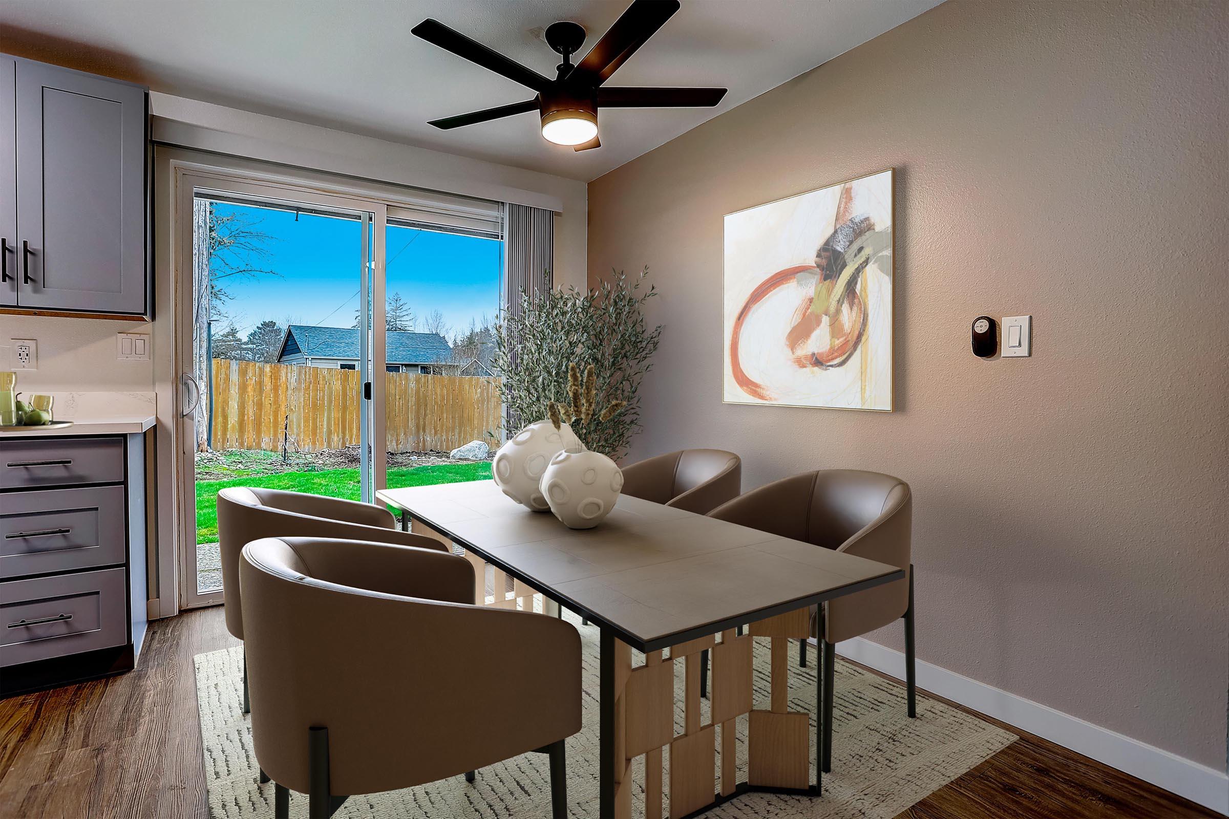 Modern dining area featuring a sleek table with minimalist chairs, a large abstract painting on the wall, and a sliding glass door that opens to a green yard. Decorative vases sit on the table, and a ceiling fan provides a contemporary touch to the room's warm color palette.