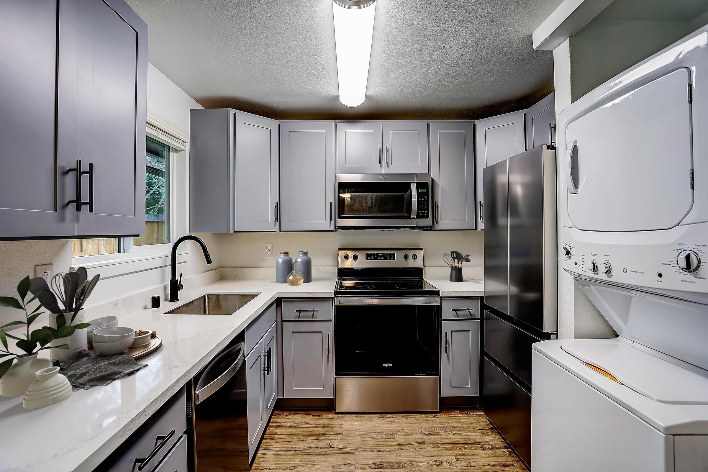 Modern kitchen with gray cabinets and white countertops. Features include a stainless steel oven, microwave, and refrigerator. A washing machine and dryer are integrated into the space. The kitchen is well-lit with a fluorescent light and has a minimalistic design, accented by decorative items on the countertop.