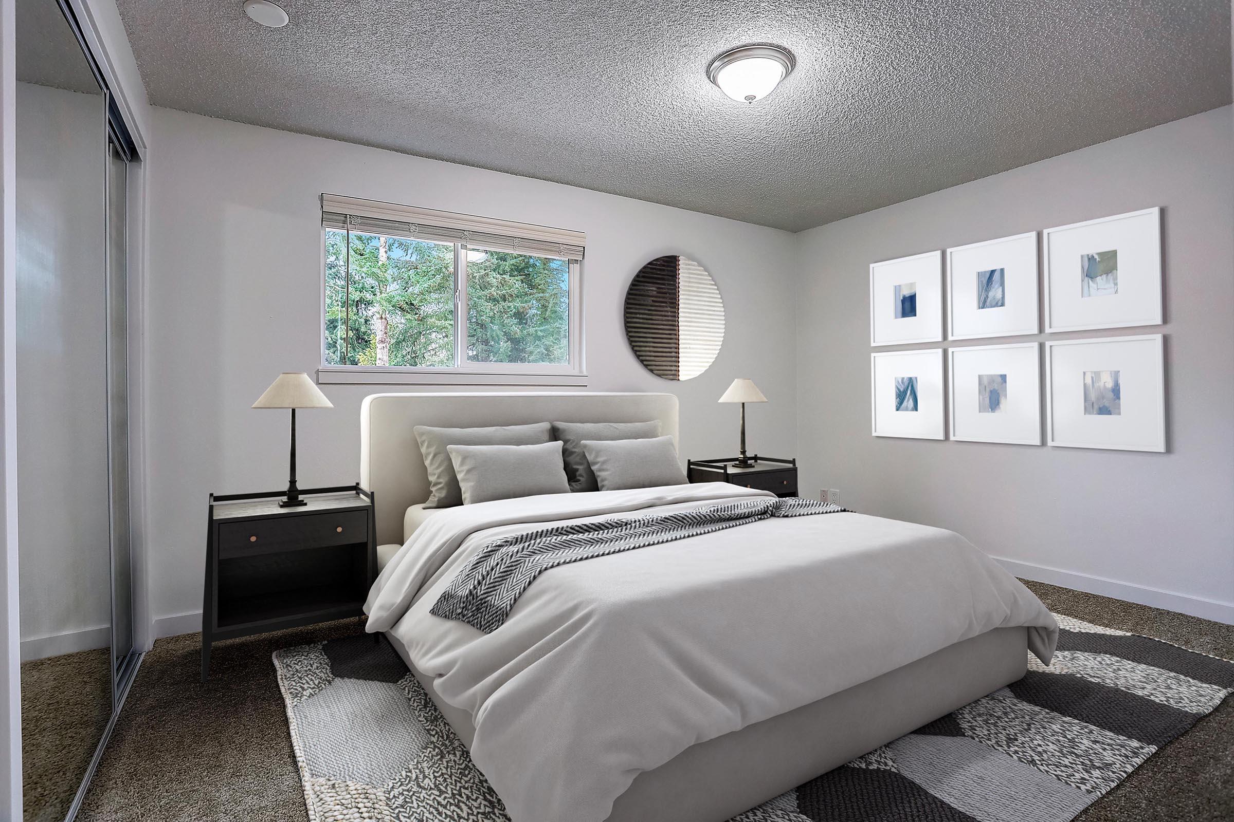 A modern bedroom featuring a queen-sized bed with light gray bedding, two bedside tables with lamps, and framed artwork on the wall. A large window allows natural light in and offers a view of greenery outside. The room has textured walls and a soft rug on the floor.