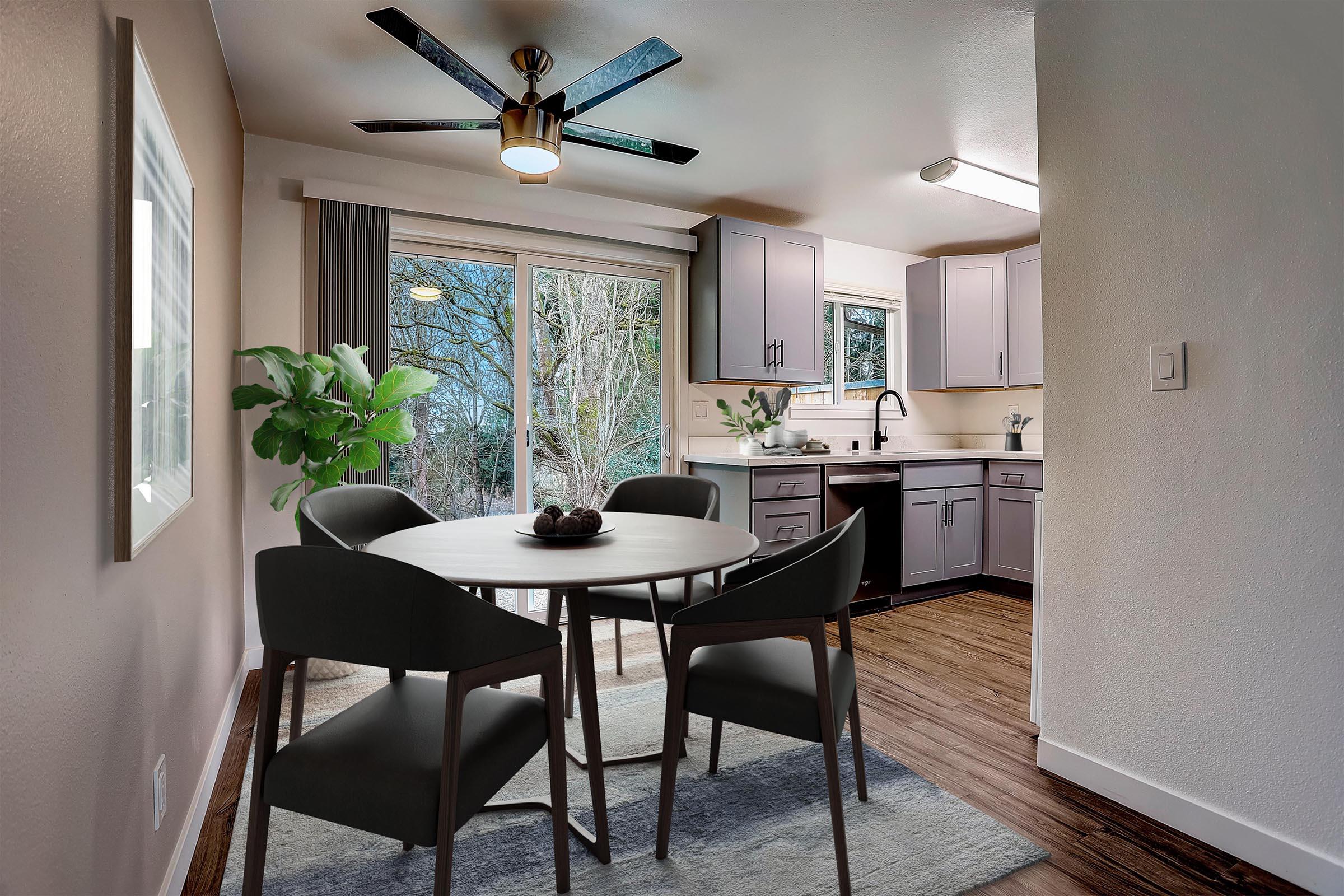 Modern kitchen and dining area featuring a round dining table with four chairs, a ceiling fan, and large sliding glass doors leading to a view of greenery outside. The kitchen has gray cabinets, white countertops, and a sink, complemented by a cozy rug under the table.