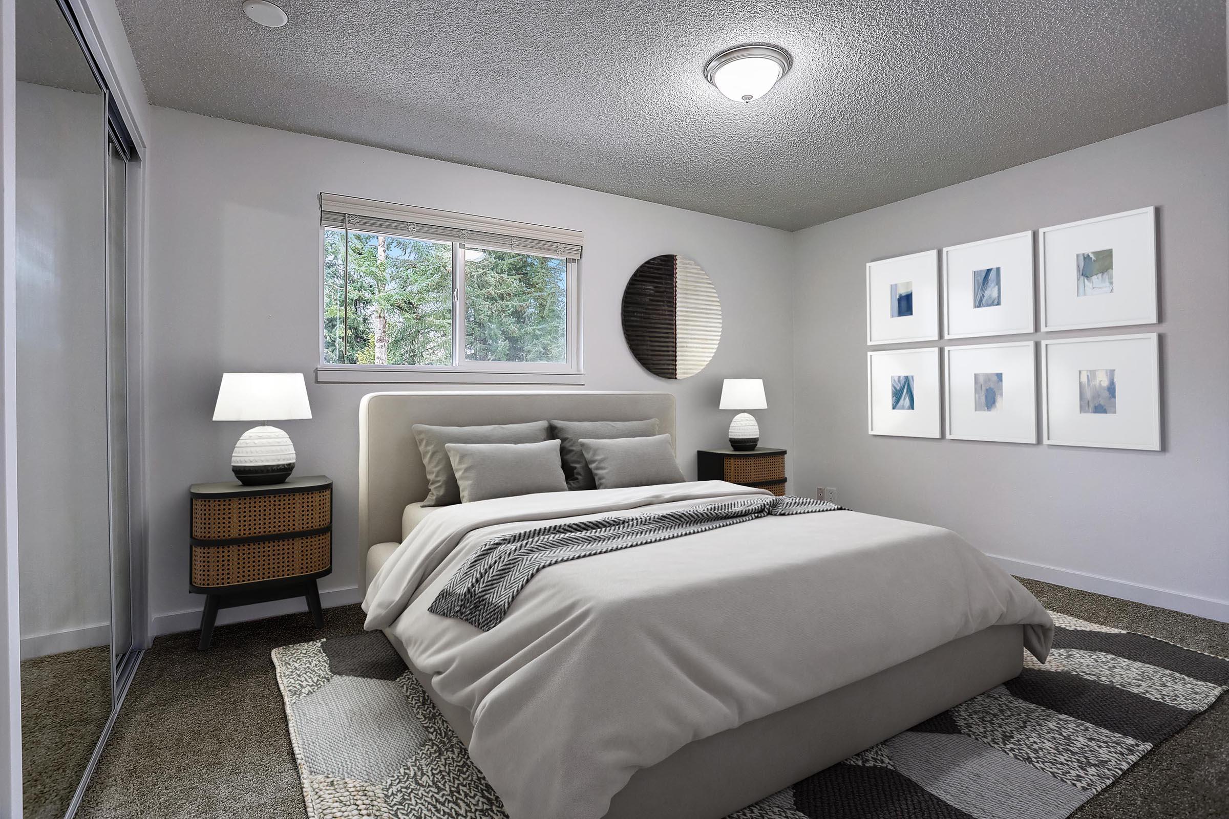 A modern bedroom featuring a light-colored upholstered bed with gray bedding. Two nightstands with lamps flank the bed. A large window provides natural light, revealing greenery outside. The walls are adorned with art, and a round mirror complements the decor, while a textured area rug adds warmth to the space.