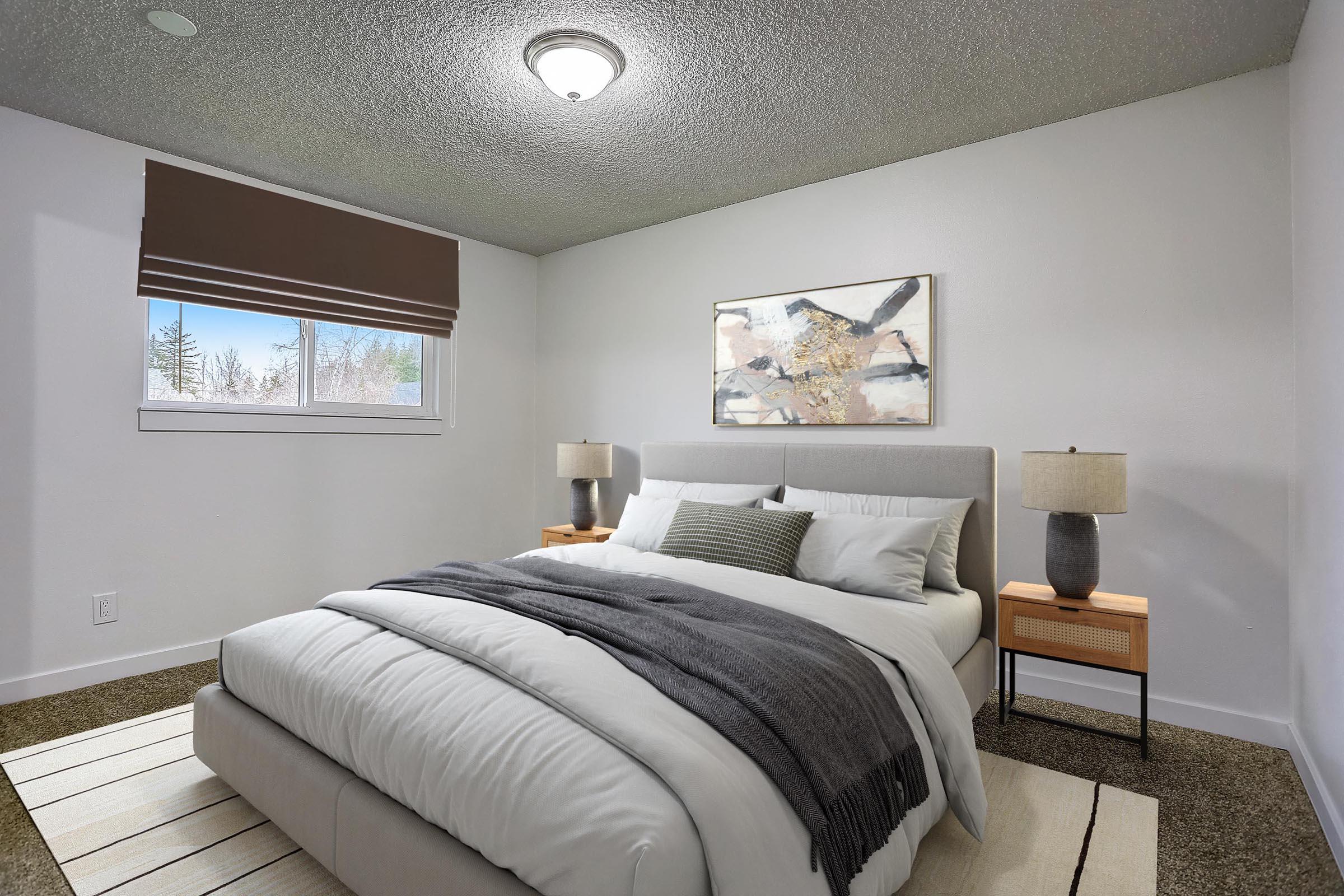 A modern bedroom featuring a queen-sized bed with white bedding and gray throw blankets. Two bedside tables with lamps flank the bed. A large window with a brown roller shade provides natural light, and a piece of abstract artwork hangs on the wall. The room has a cozy, contemporary aesthetic.