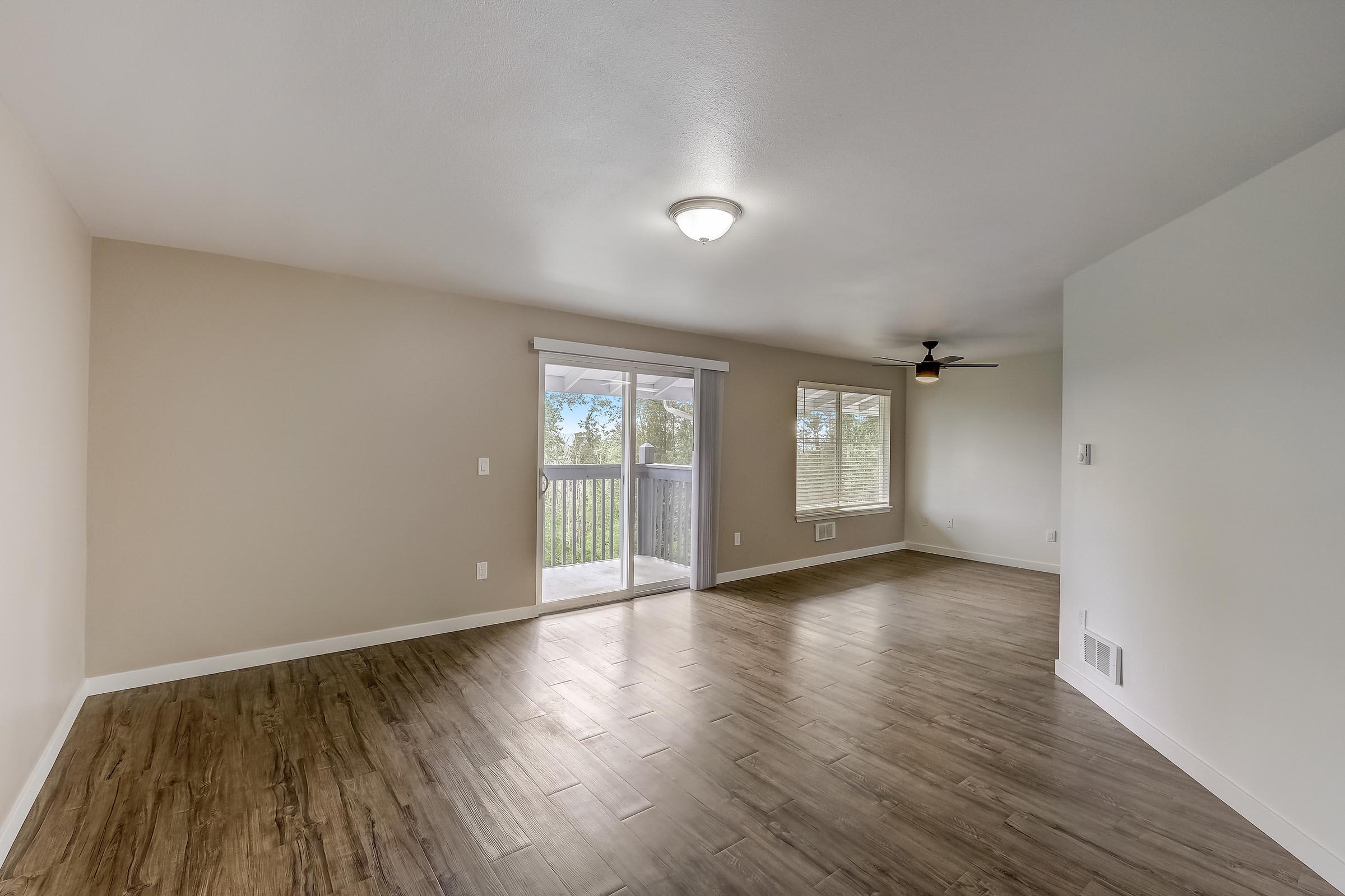 Spacious, sunlit living room featuring light-colored walls, hardwood-style flooring, and a ceiling light fixture. Large sliding glass doors lead to a balcony with scenic views, and several windows provide natural light. The room is empty, offering a blank canvas for decorating.