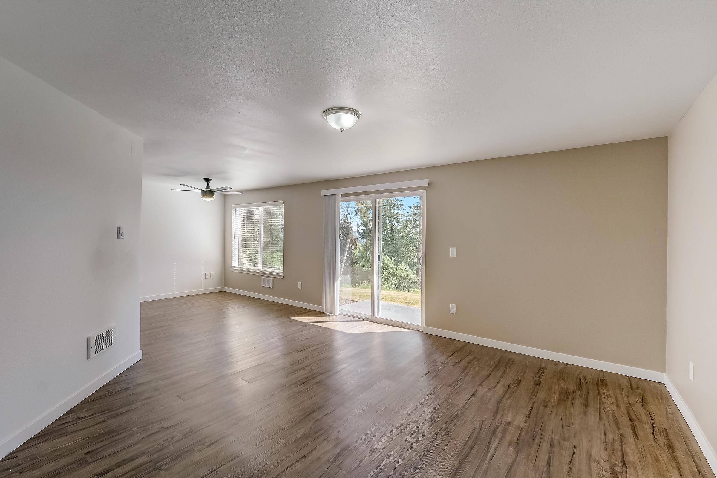 Empty room with light brown walls and a large sliding glass door leading to a patio. The floor is finished with light wood-like flooring. A ceiling fan is visible, and there are windows allowing natural light. The space is bright and unembellished, suggesting potential for personalization.