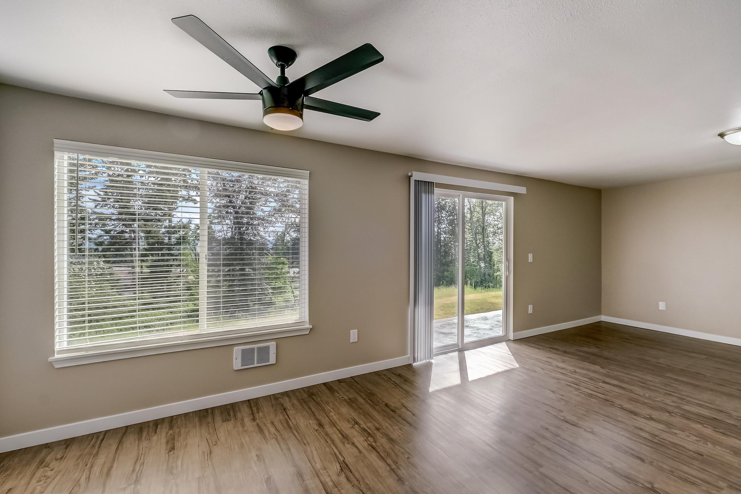 Bright, open interior of a room with light-colored walls and wooden flooring. A ceiling fan is installed, and large windows allow natural light to enter. Sliding glass doors lead to an outdoor area, with greenery visible through the windows. The space is uncluttered and inviting.