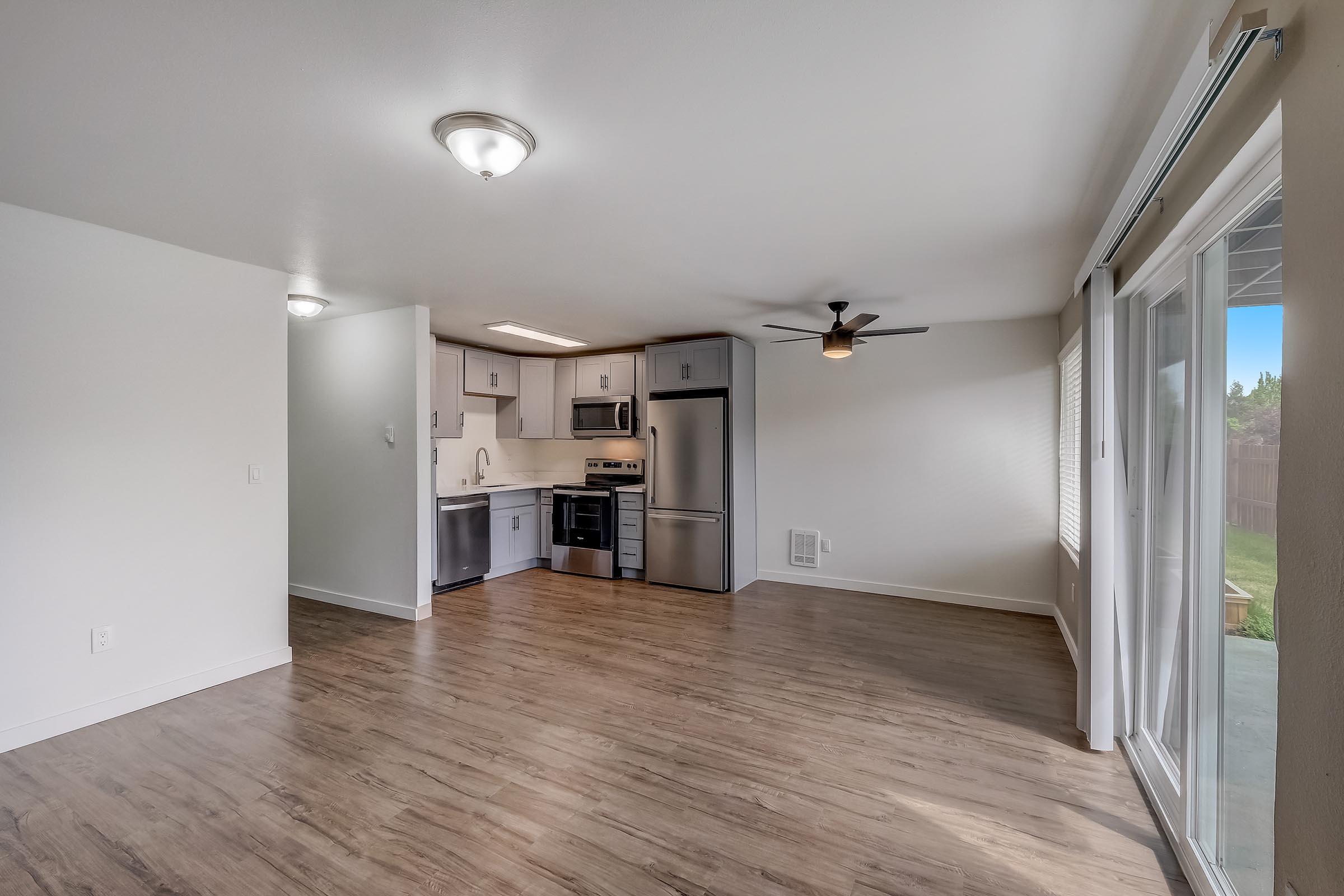 Bright, modern interior of an open-concept living space featuring light-colored walls, hardwood flooring, and a kitchen area with stainless steel appliances. A ceiling fan is visible, and sliding glass doors lead to an outdoor space. The layout is spacious and inviting, perfect for comfortable living.