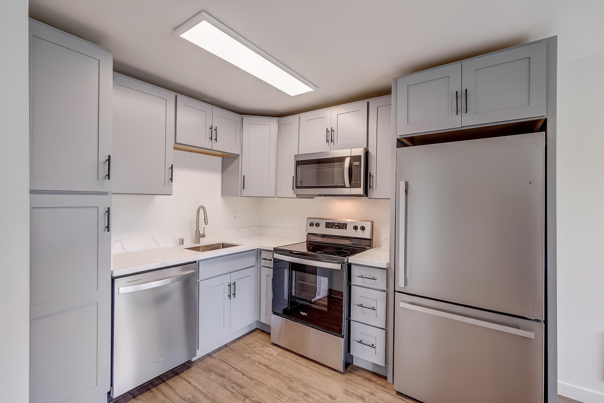 Modern kitchen with light gray cabinets, stainless steel appliances, a built-in microwave, and a sleek oven. Features a double sink and a large refrigerator. The space has a clean, contemporary design with warm wood-like flooring and ample natural light from the overhead fixture.