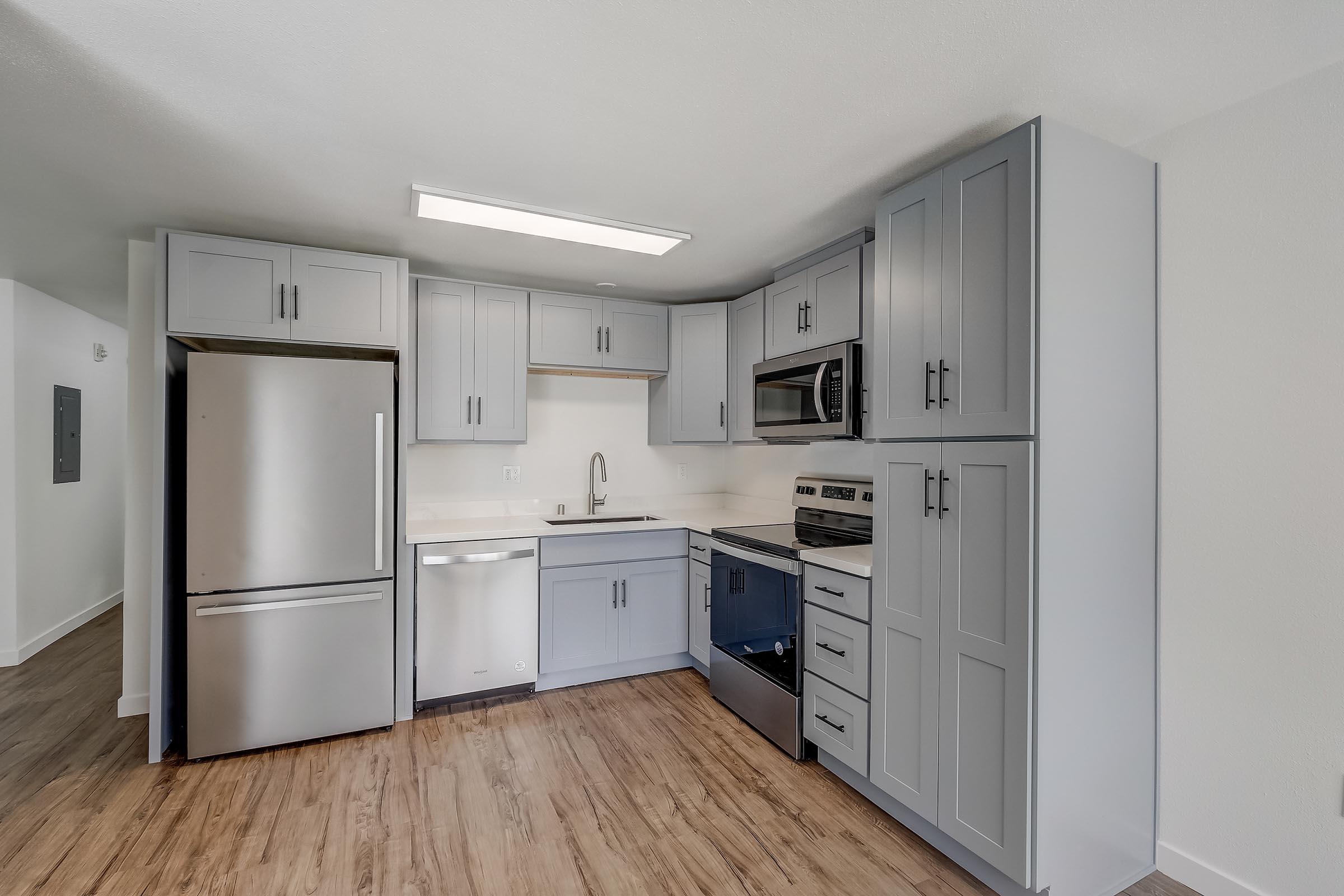 A modern kitchen featuring gray cabinetry, stainless steel appliances including a fridge and oven, a microwave, and a sink. The kitchen has a light-colored countertop and a spacious layout with wood-like flooring, creating a contemporary and inviting atmosphere.